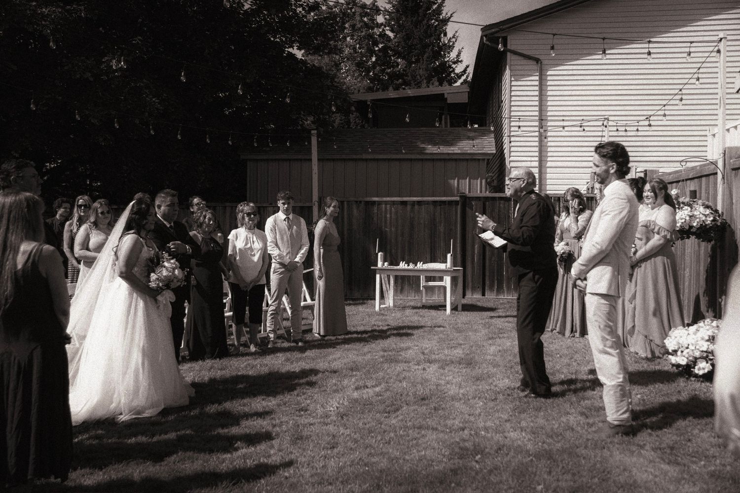 Black and white outdoor wedding ceremony in backyard with bride, groom, officiant, and guests gathered on lawn.