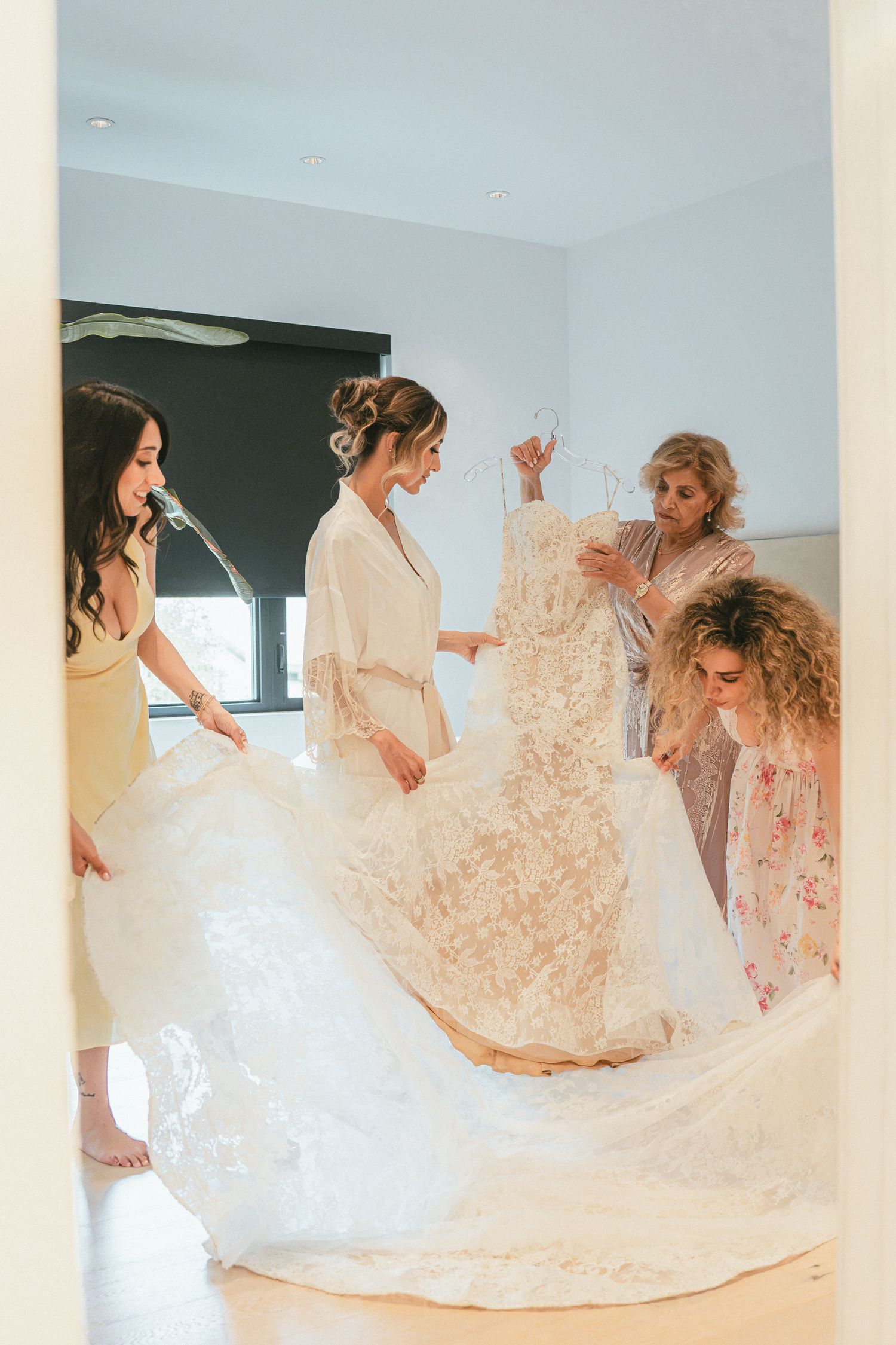 Bridesmaids helping bride into a stunning lace wedding gown with a long train in a bright room.