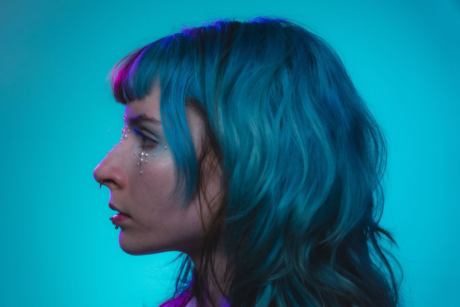 Side profile of woman with teal hair and pearl face gems against a bright turquoise background.