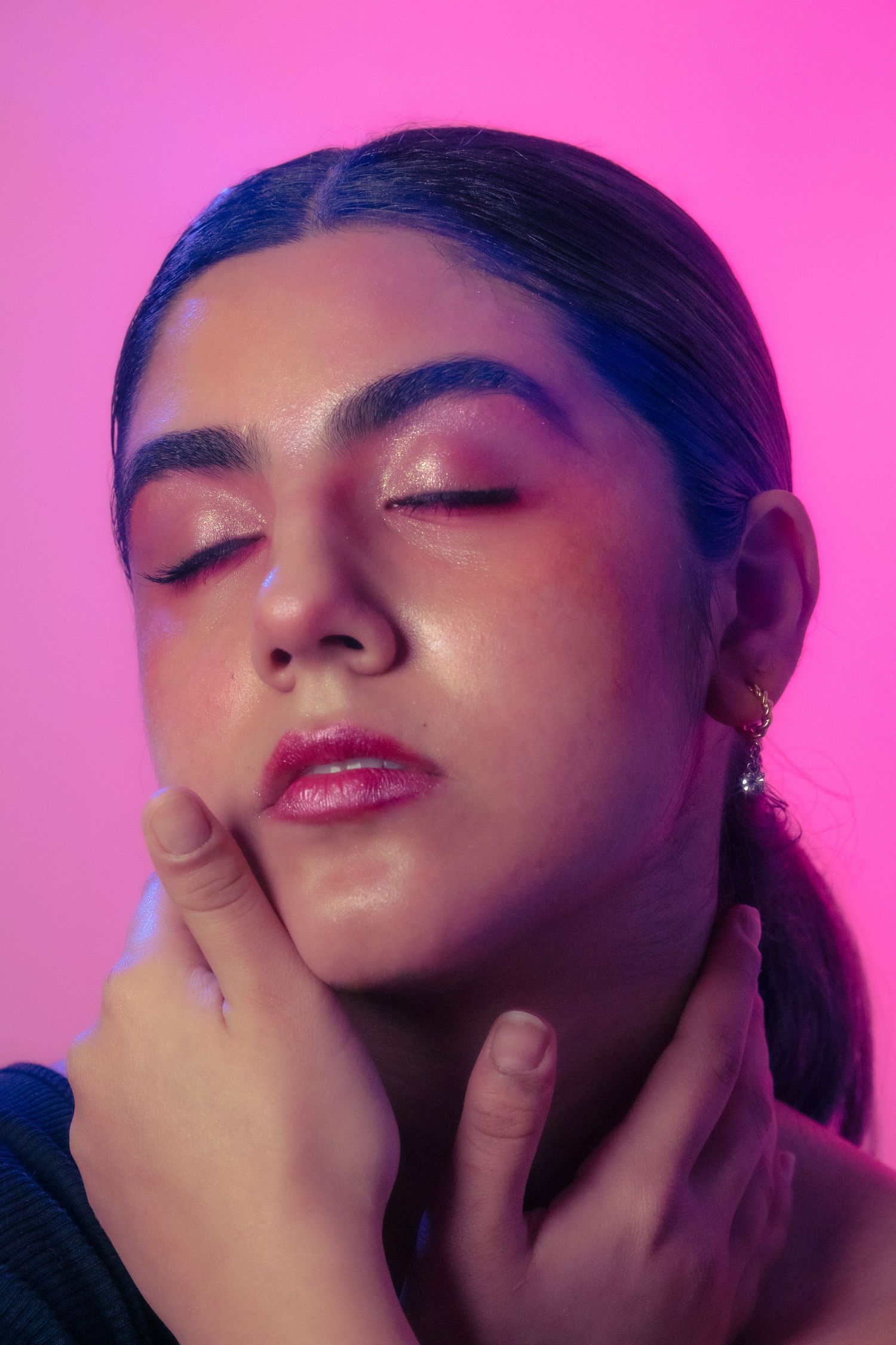 Beauty portrait of a woman with bold pink eye makeup touching her face against a neon pink background.