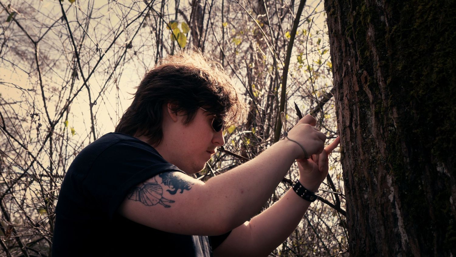 Tattooed person examining tree bark closely in a woodland setting.