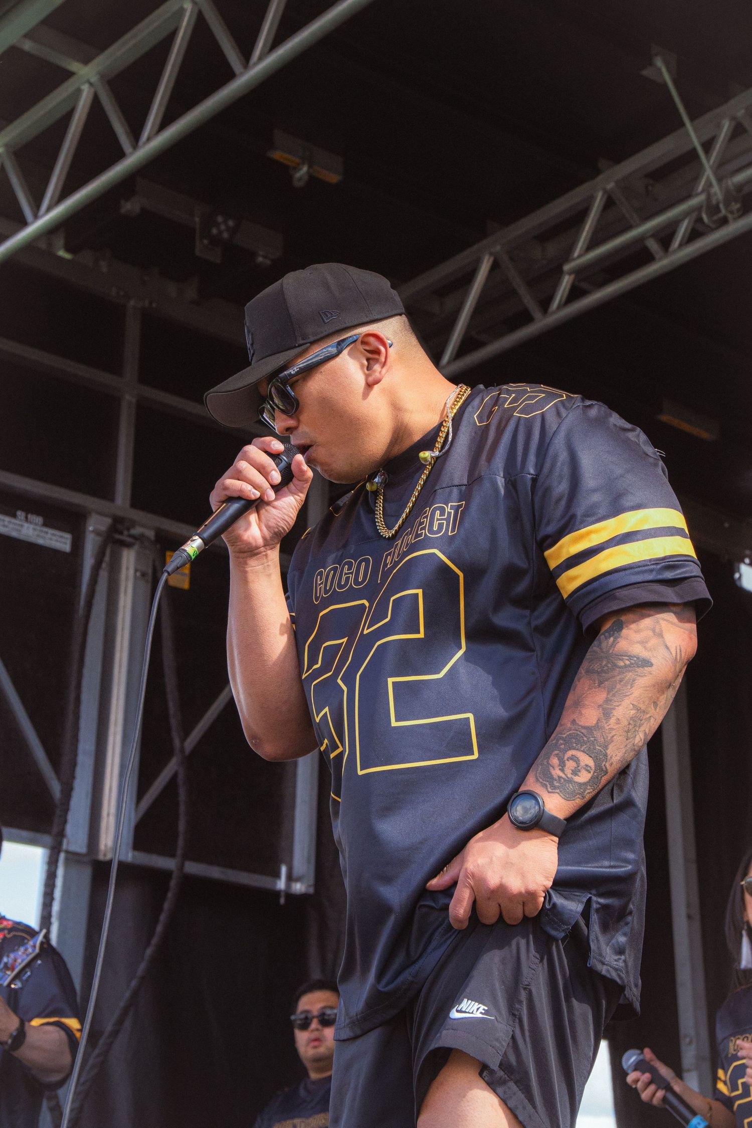 Male performer in Coco Project jersey and cap leans forward singing into microphone on stage.
