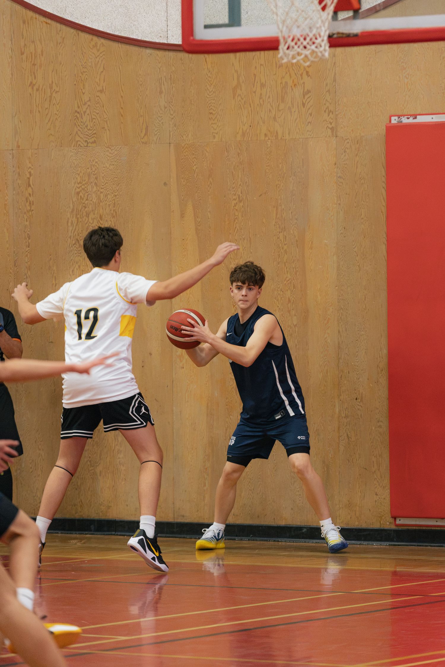 Youth basketball player in navy uniform holds ball while being defended by opponent #12 in white jersey indoors.