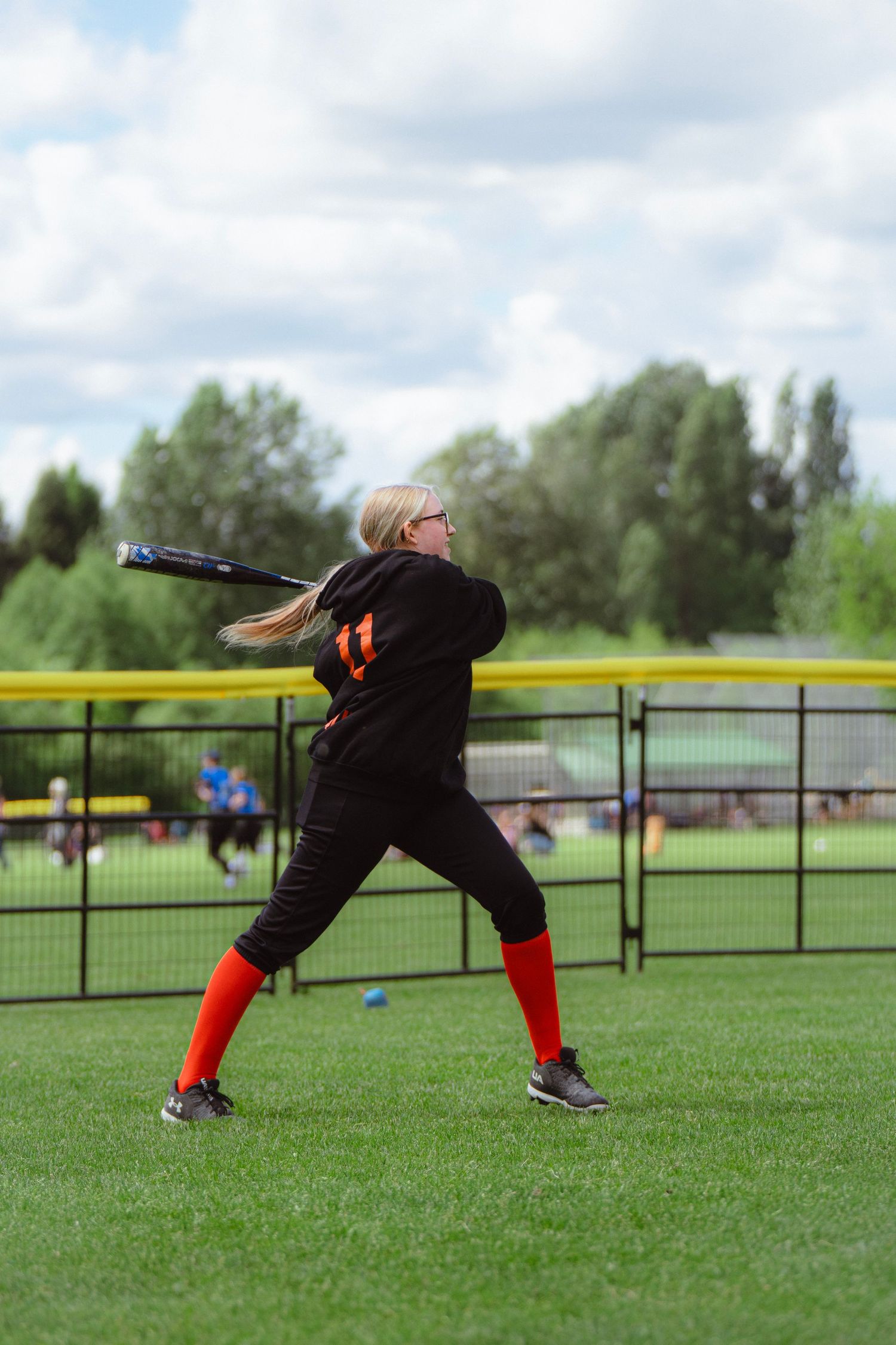 Blonde female softball player in black hoodie swinging bat on field.
