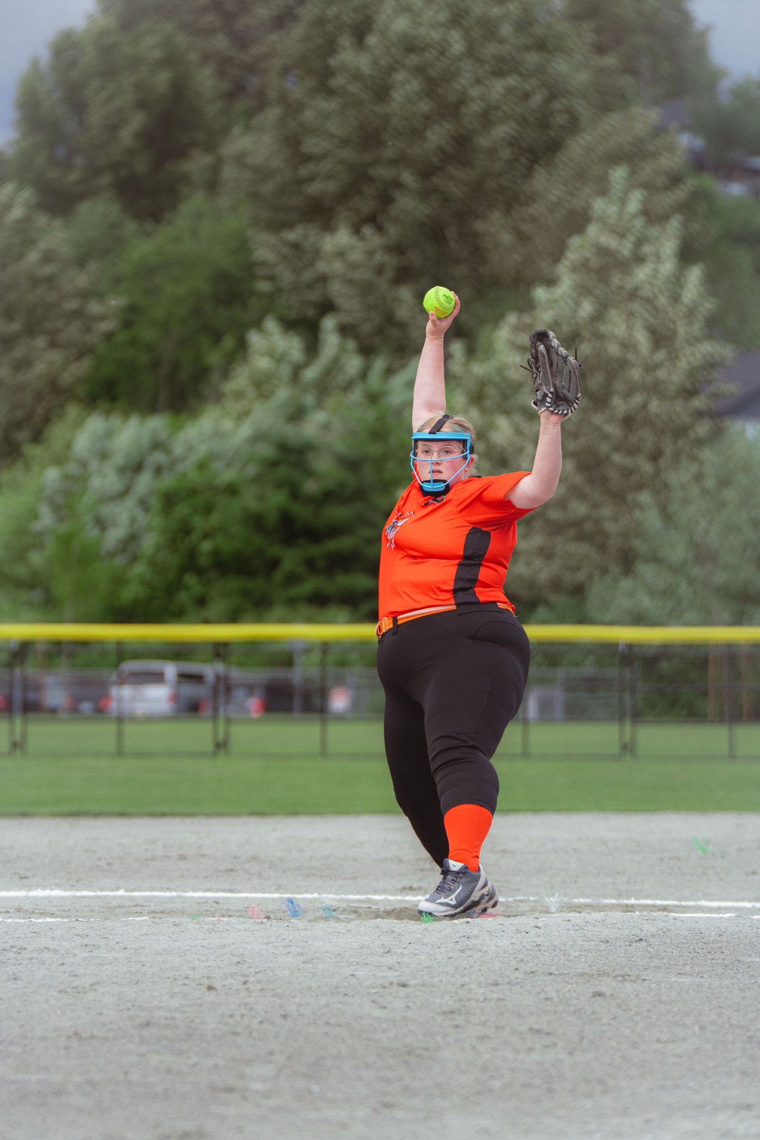 Female softball pitcher in orange jersey delivers windmill pitch on overcast day at outdoor diamond.