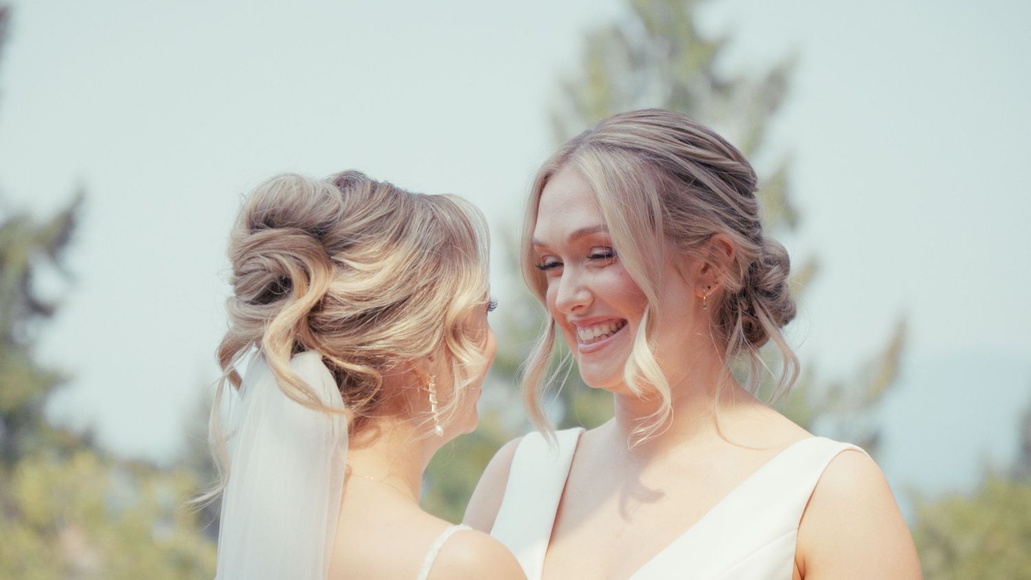 Two smiling bridesmaids in white dresses with elegant updo hairstyles at an outdoor wedding celebration.