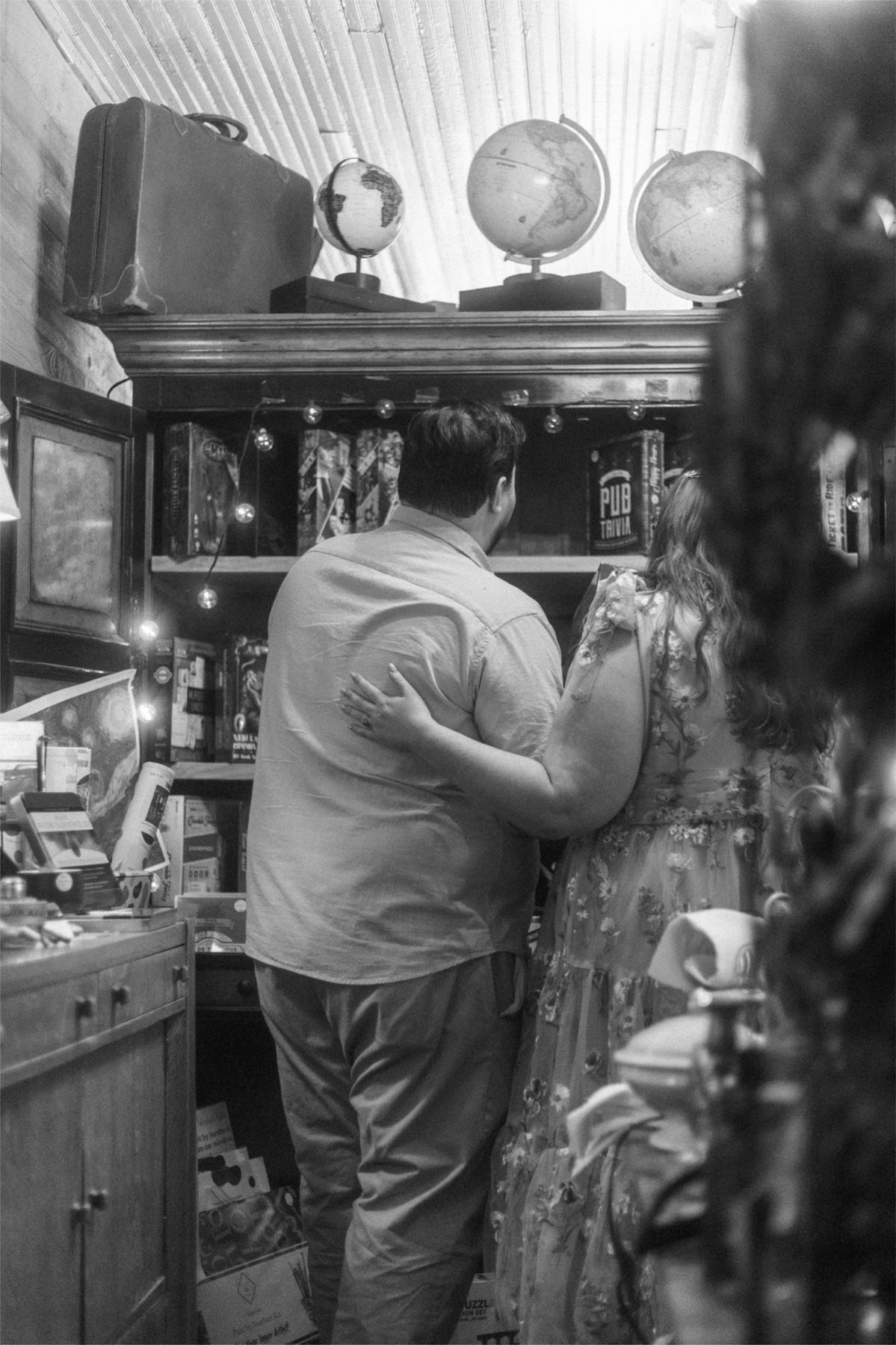 Black and white photo of a couple embracing near a bookshelf decorated with vintage globes and suitcases.