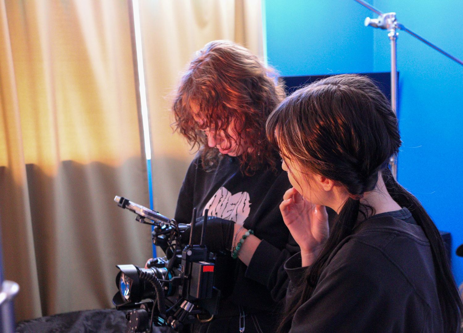 Two filmmakers operate a professional video camera together on an indoor production set.