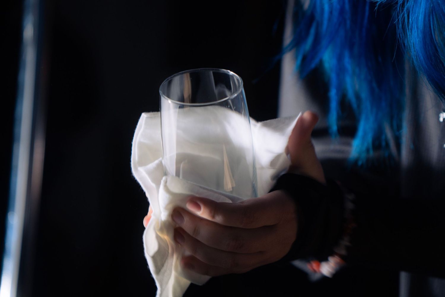 Person with blue hair holding a clear glass wrapped in white fabric against a dark background.