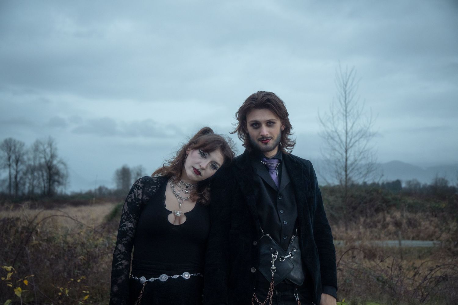 Gothic couple in black clothing posing in an overcast outdoor field setting.