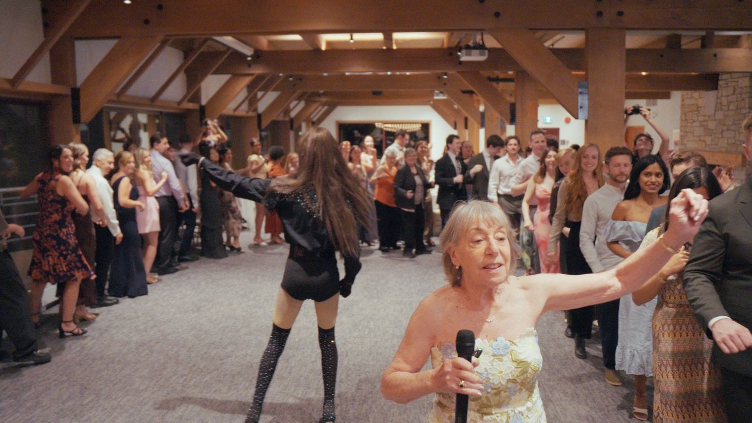 A wedding entertainer in black interacts with a bride holding a microphone before a large crowd.