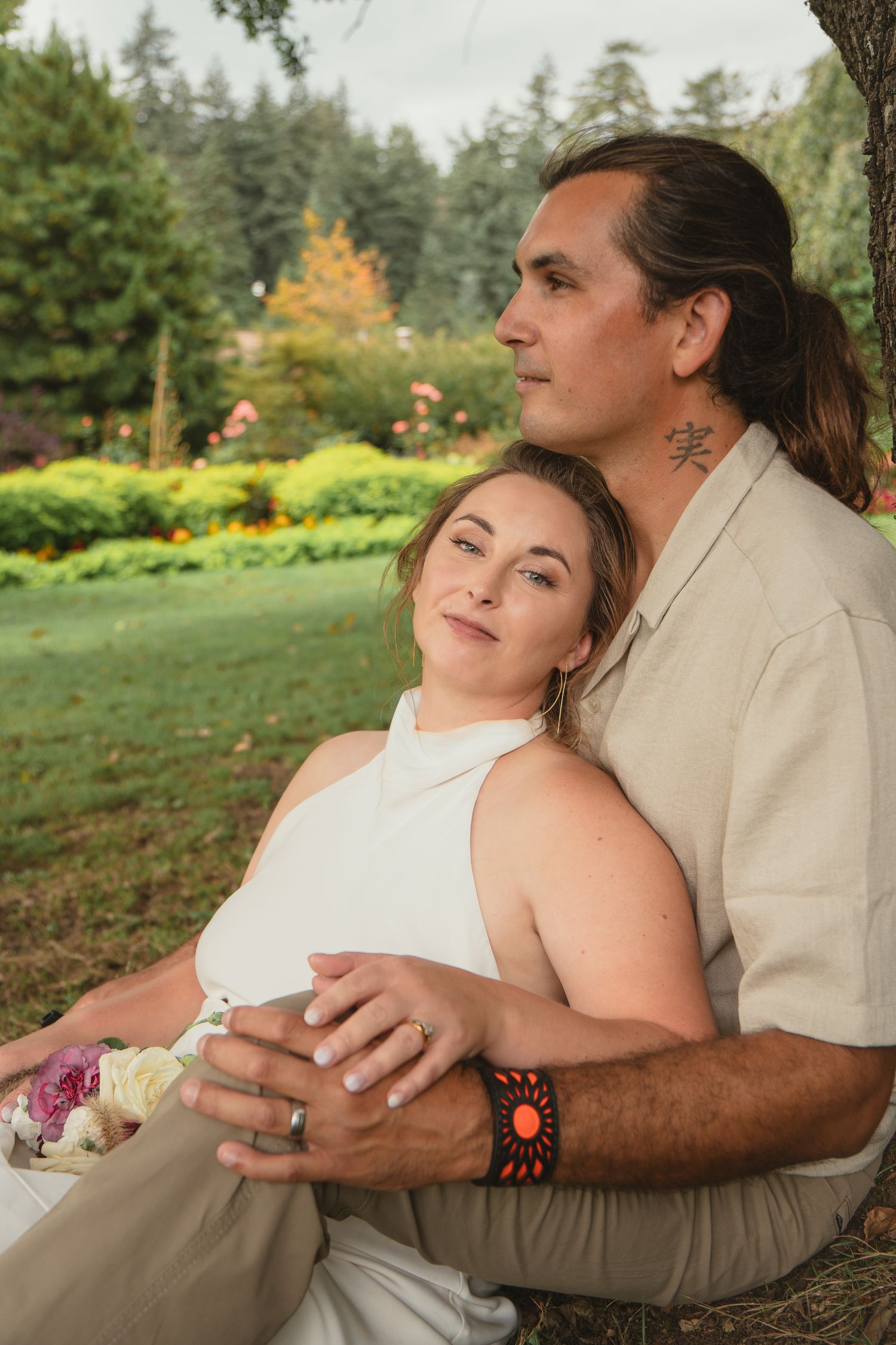 Bride rests against groom beneath a tree as they gaze peacefully at a colorful garden landscape.