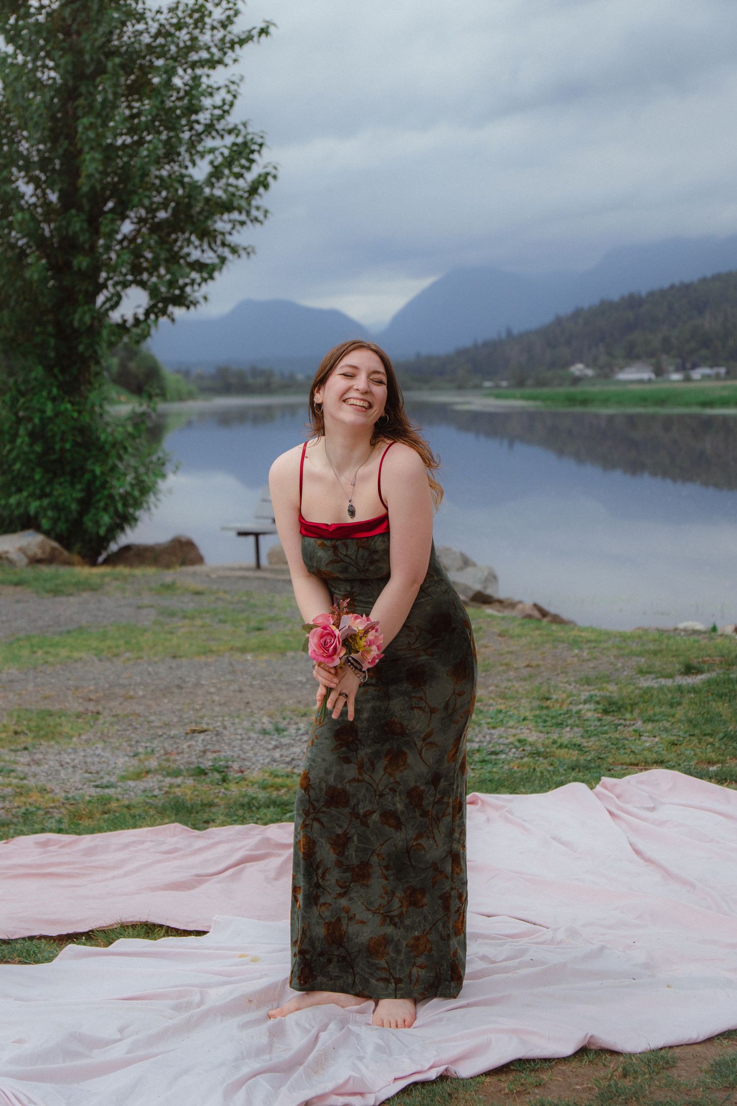 Laughing woman in dark floral maxi dress holds pink roses by a misty mountain lake.