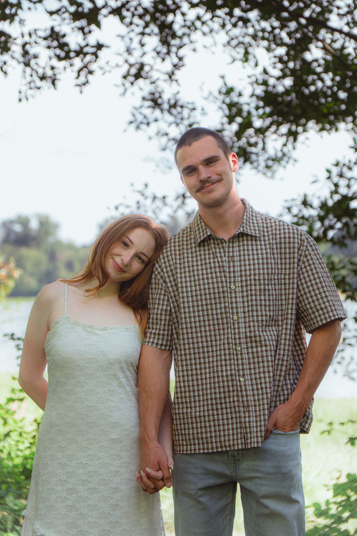 A young couple holds hands and poses together near a serene lake surrounded by lush green trees.