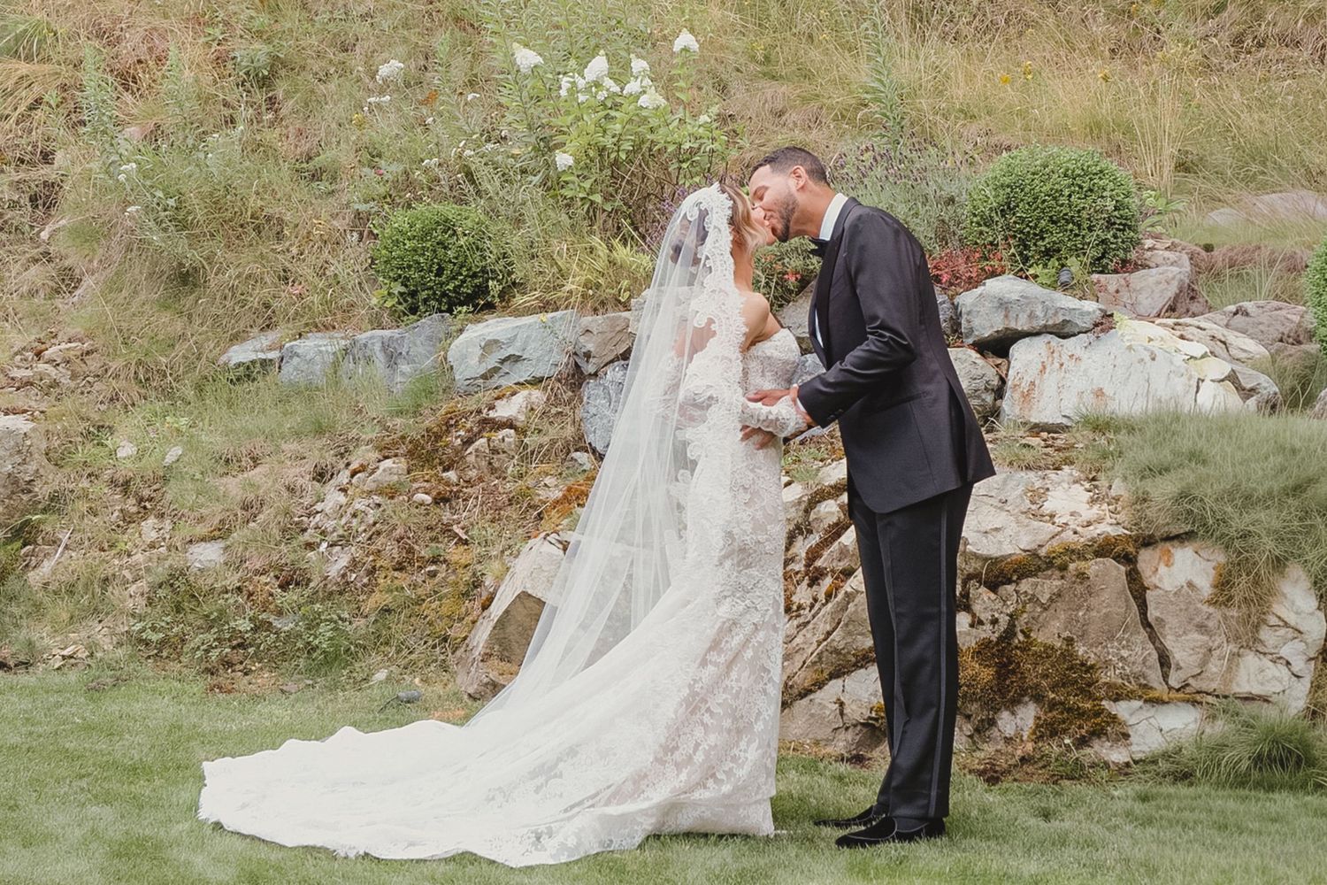 Bride and groom share a romantic kiss outdoors, with bride in lace gown and veil, groom in black tuxedo.