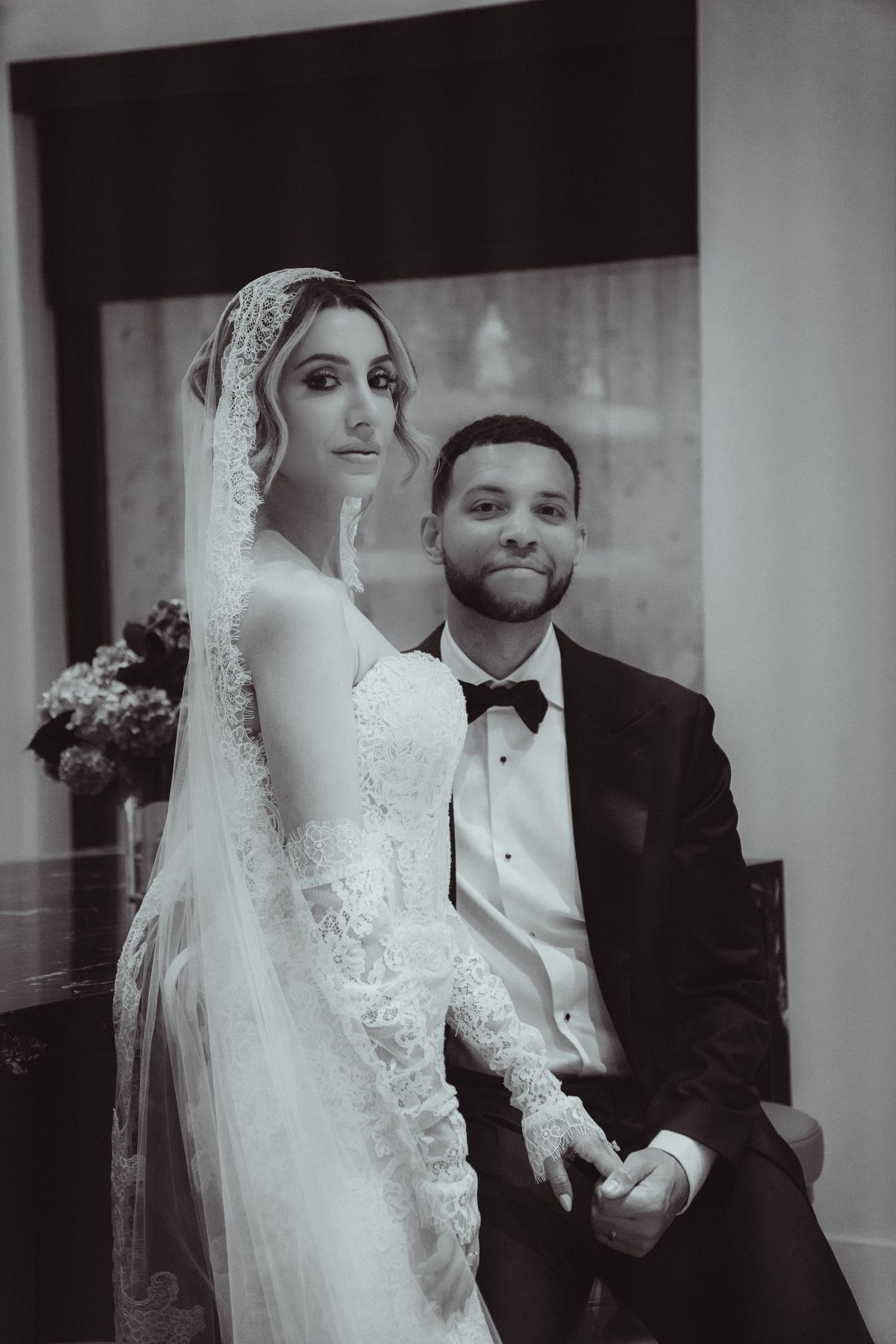 Black and white wedding photo of bride in lace gown and veil with groom in tuxedo posing elegantly together.