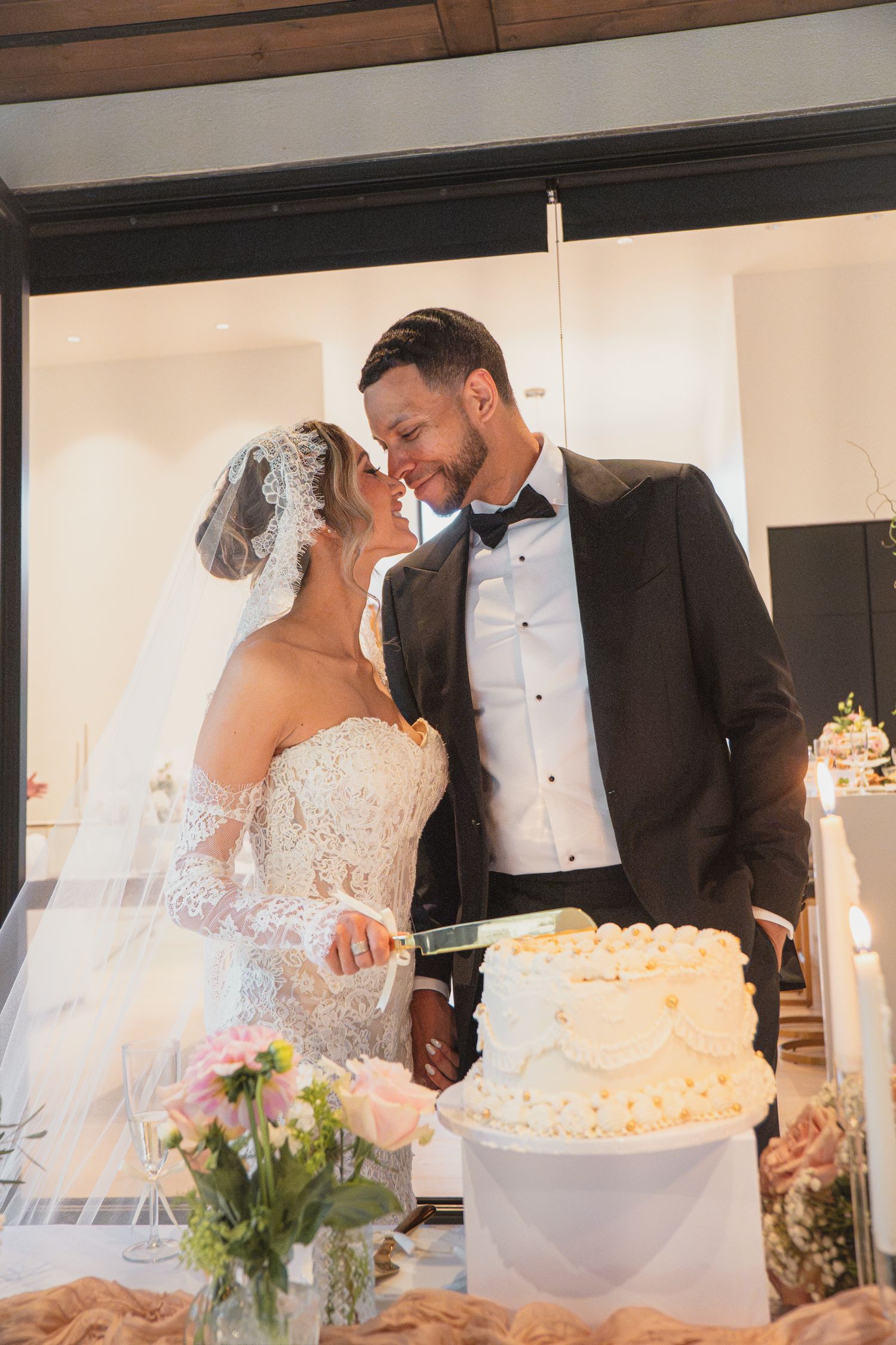 Bride and groom in wedding attire share a kiss while cutting white tiered wedding cake at reception.