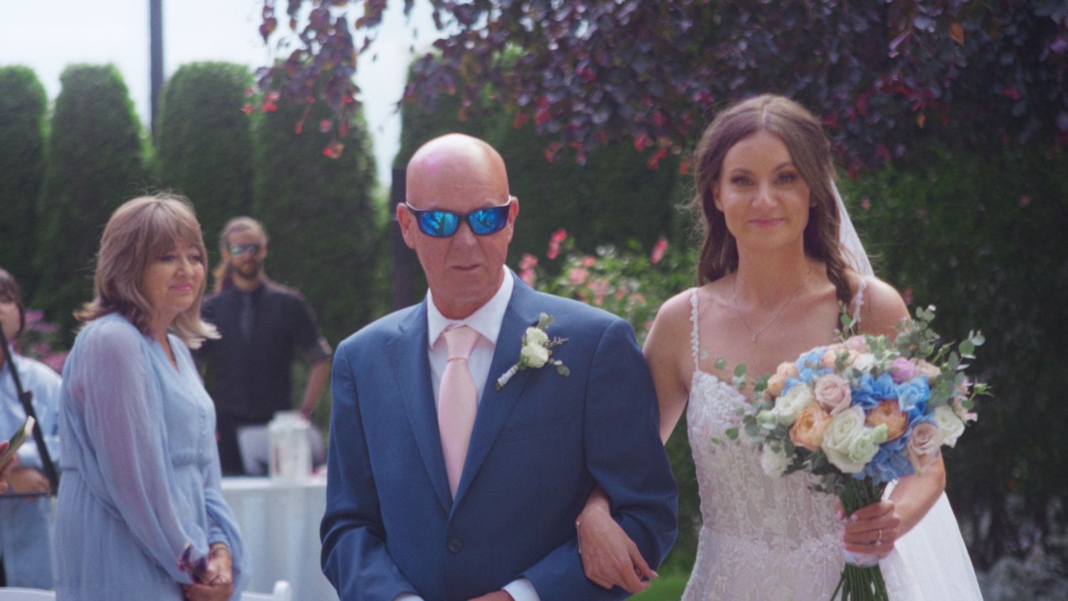 Bride walks down the aisle with her father in a blue suit during an outdoor wedding ceremony.