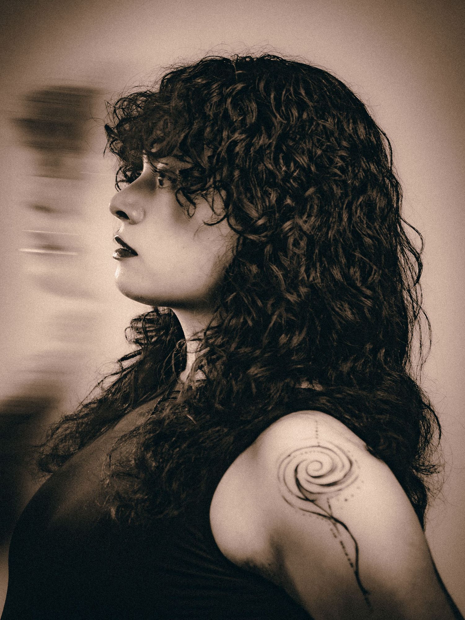 Side profile of a curly-haired woman with a spiral tattoo on her shoulder in sepia photography.