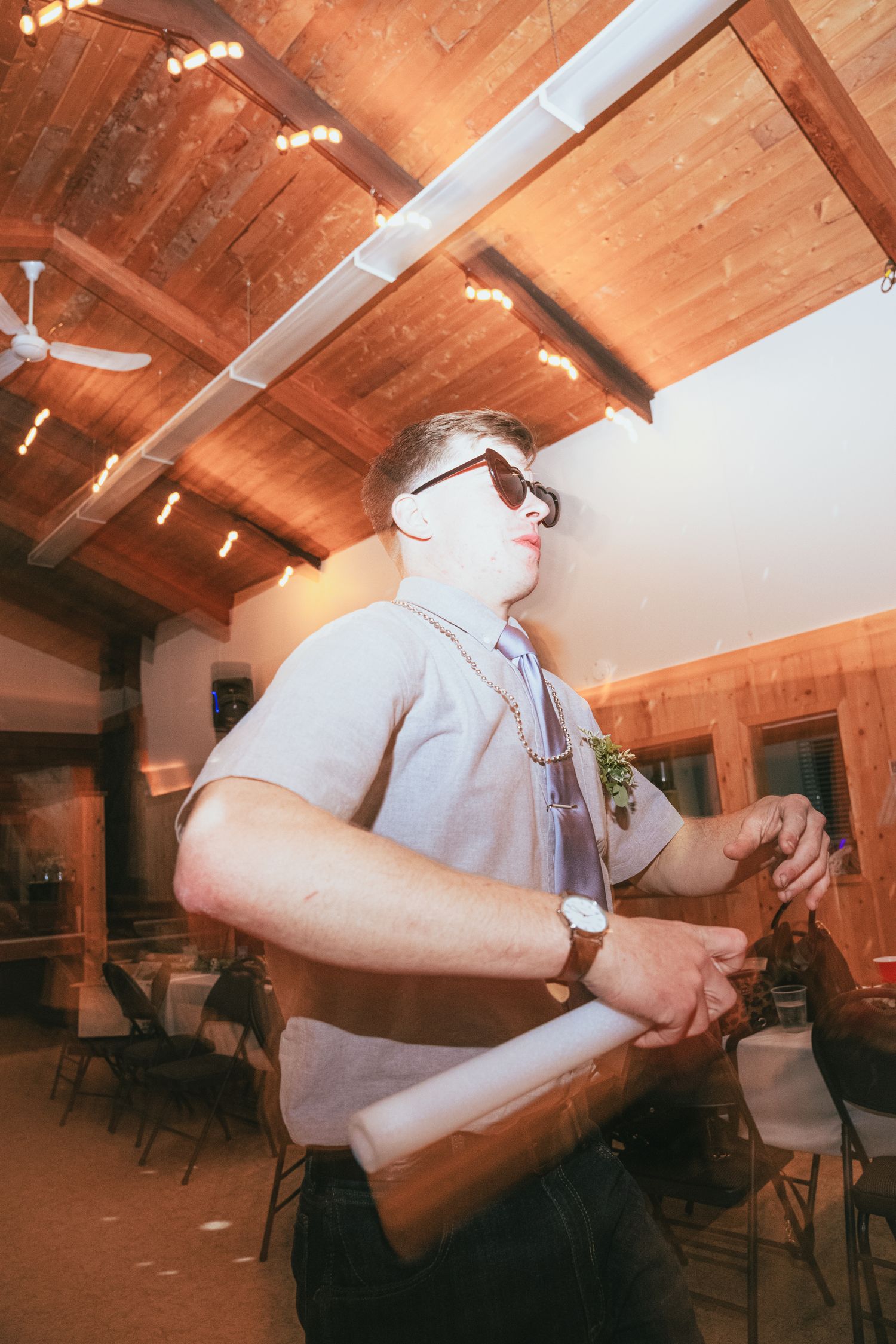 Man in sunglasses holding glow stick at warmly lit wooden wedding reception hall.