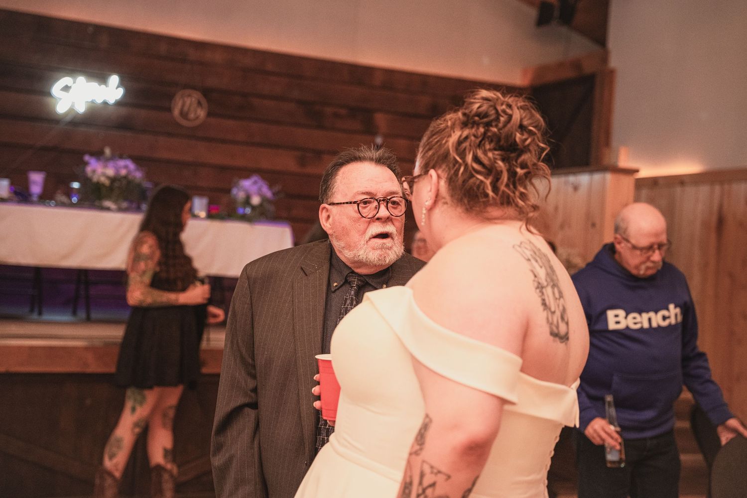 Bride with tattoos sharing a heartfelt moment with elderly guest in black suit at wedding reception.