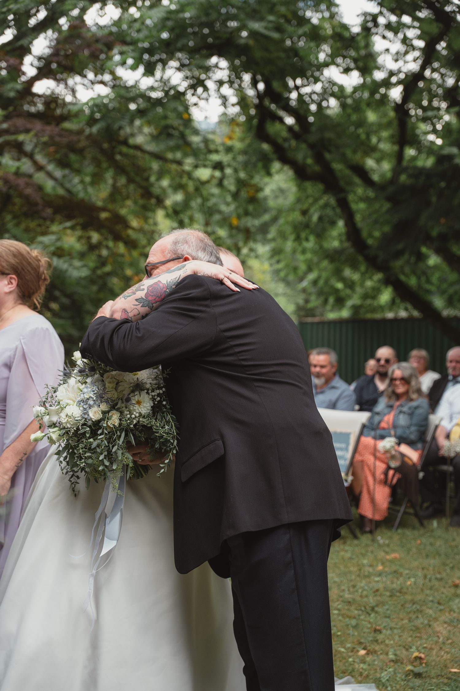 Bride in white gown embraces a man in black suit during outdoor wedding ceremony while holding bouquet.