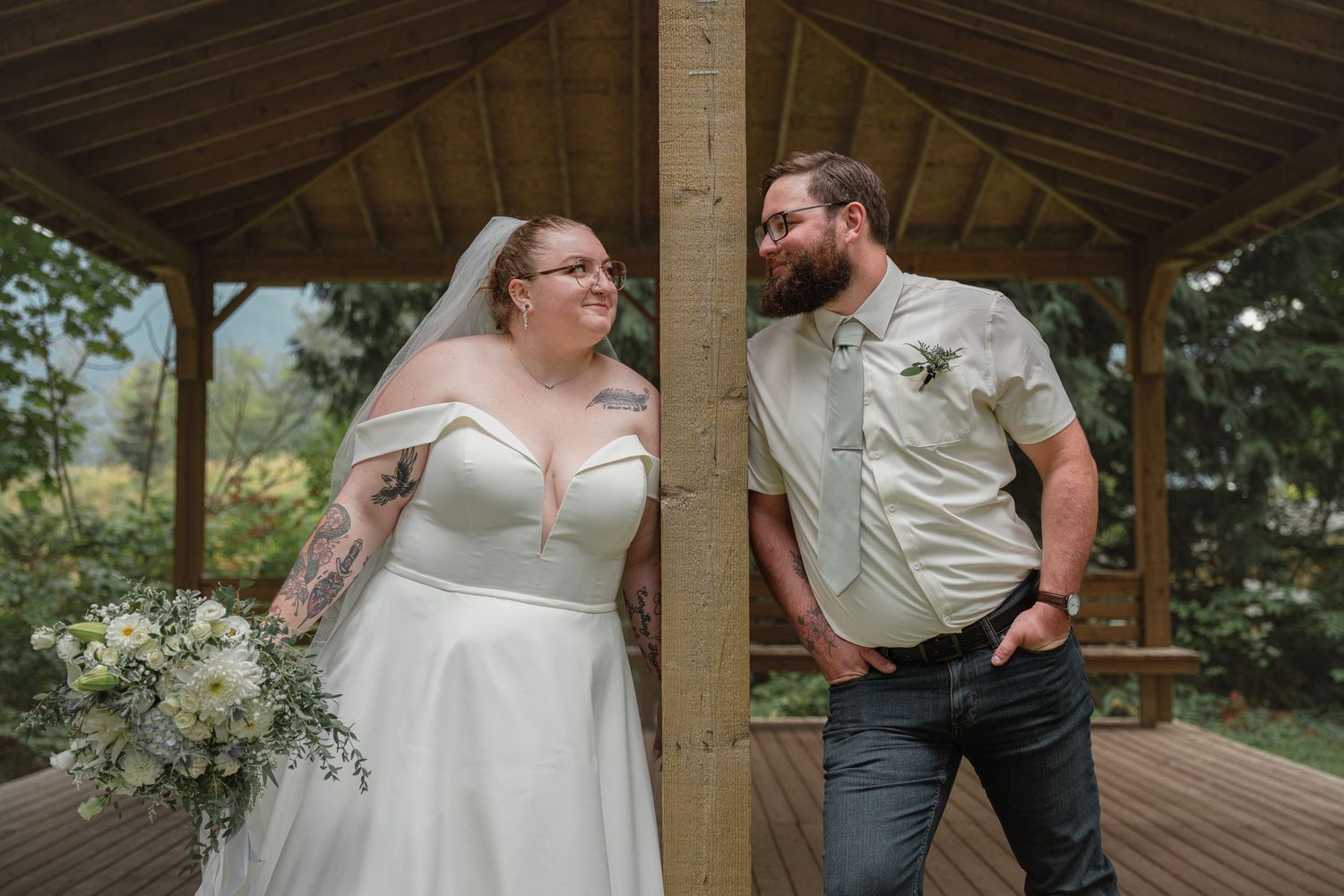 Bride in white off-shoulder gown holding bouquet smiles at bearded groom in khaki shirt under wooden gazebo.