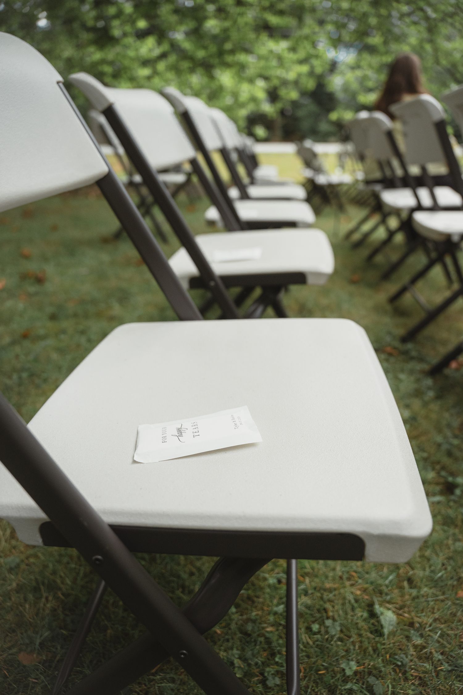 White folding chairs arranged on grass for an outdoor wedding ceremony with a printed program resting on the nearest seat.