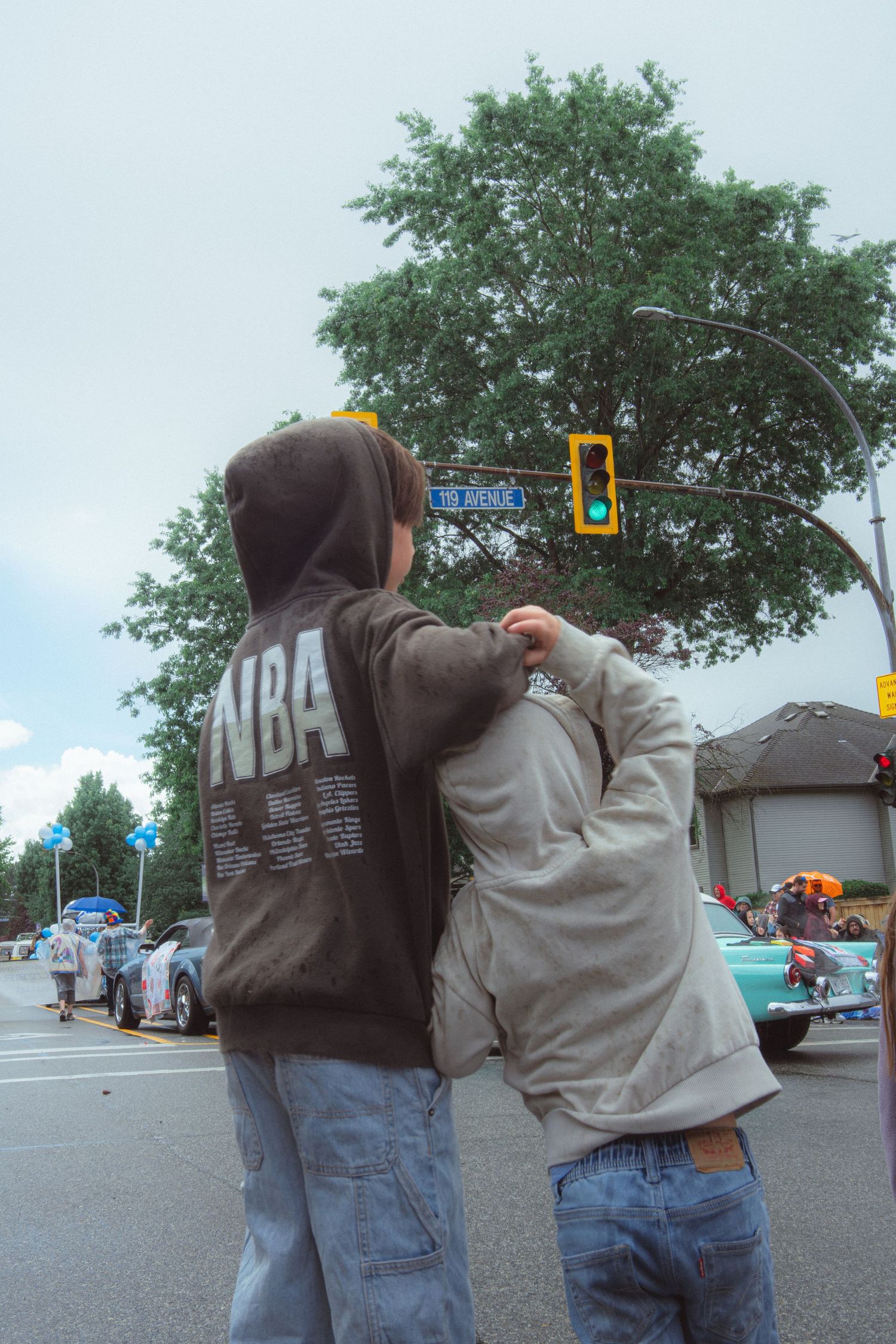 Two children in hoodies stand at a street corner watching a parade under traffic lights.