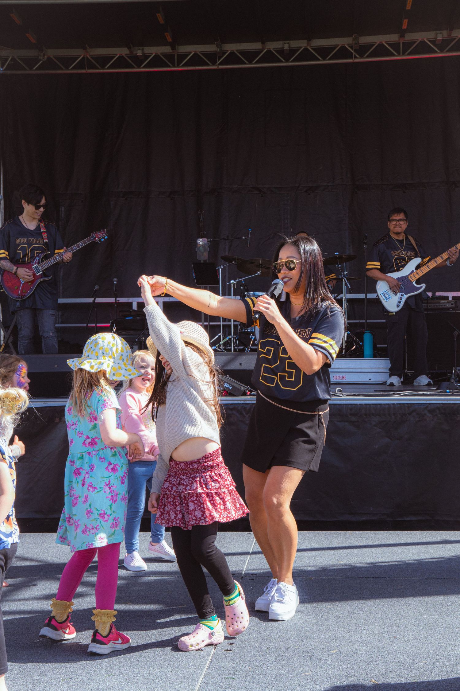 Female singer dances with young children in front of a live band on an outdoor stage at a festival.