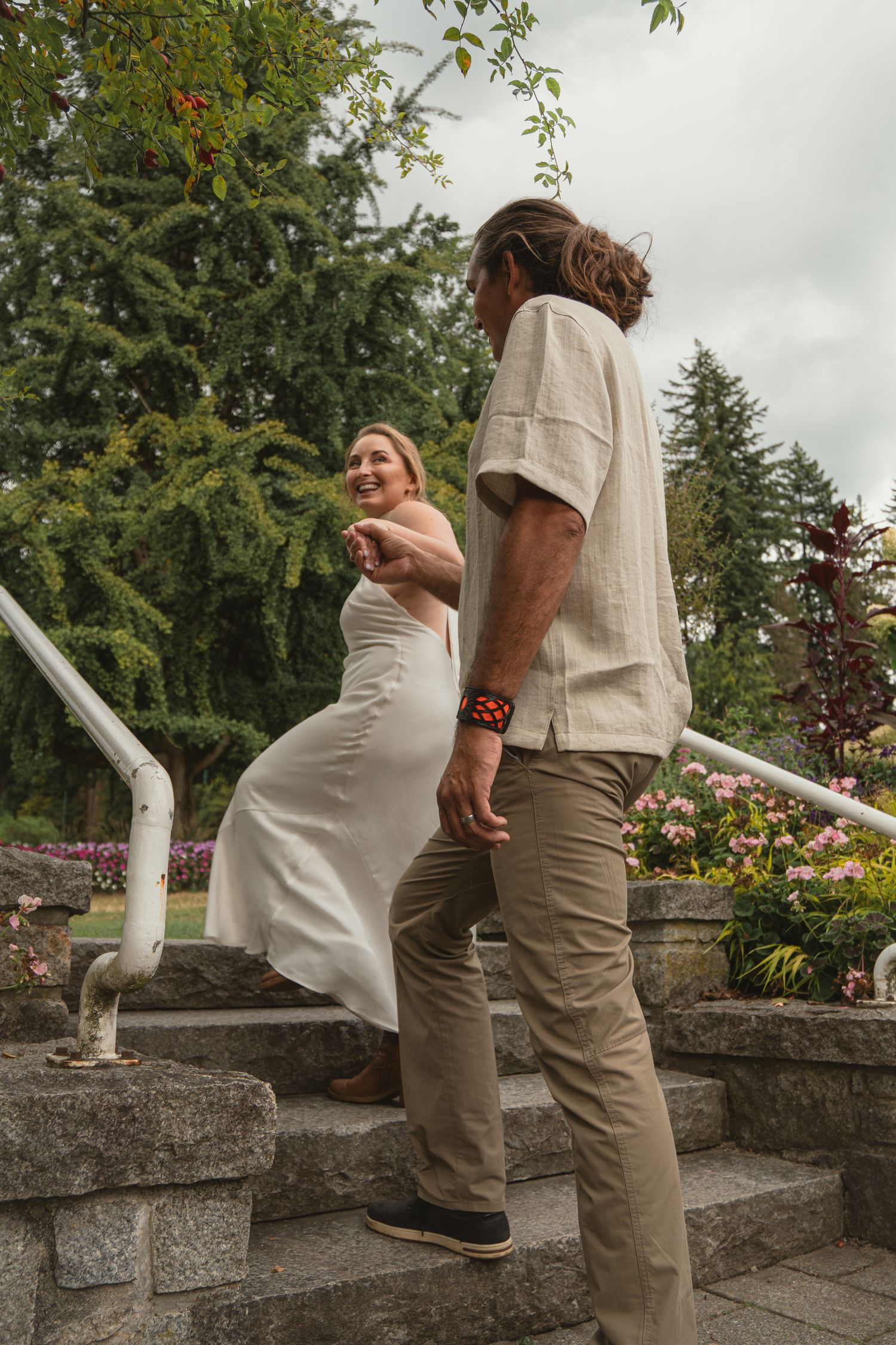 Bride in white dress leading groom up stone steps in a lush garden setting.