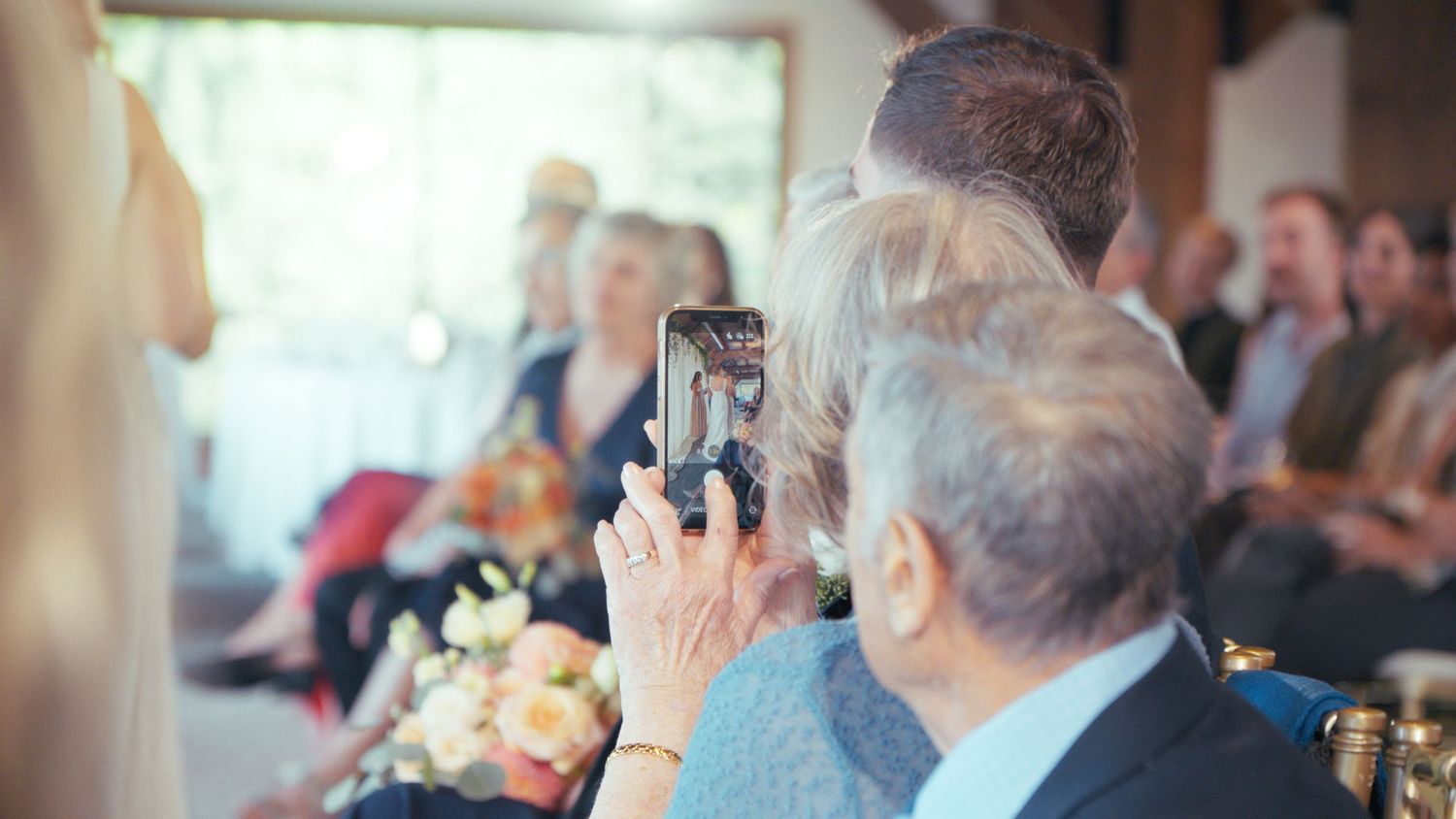 Wedding guest photographs the ceremony on a smartphone among seated guests holding flowers.