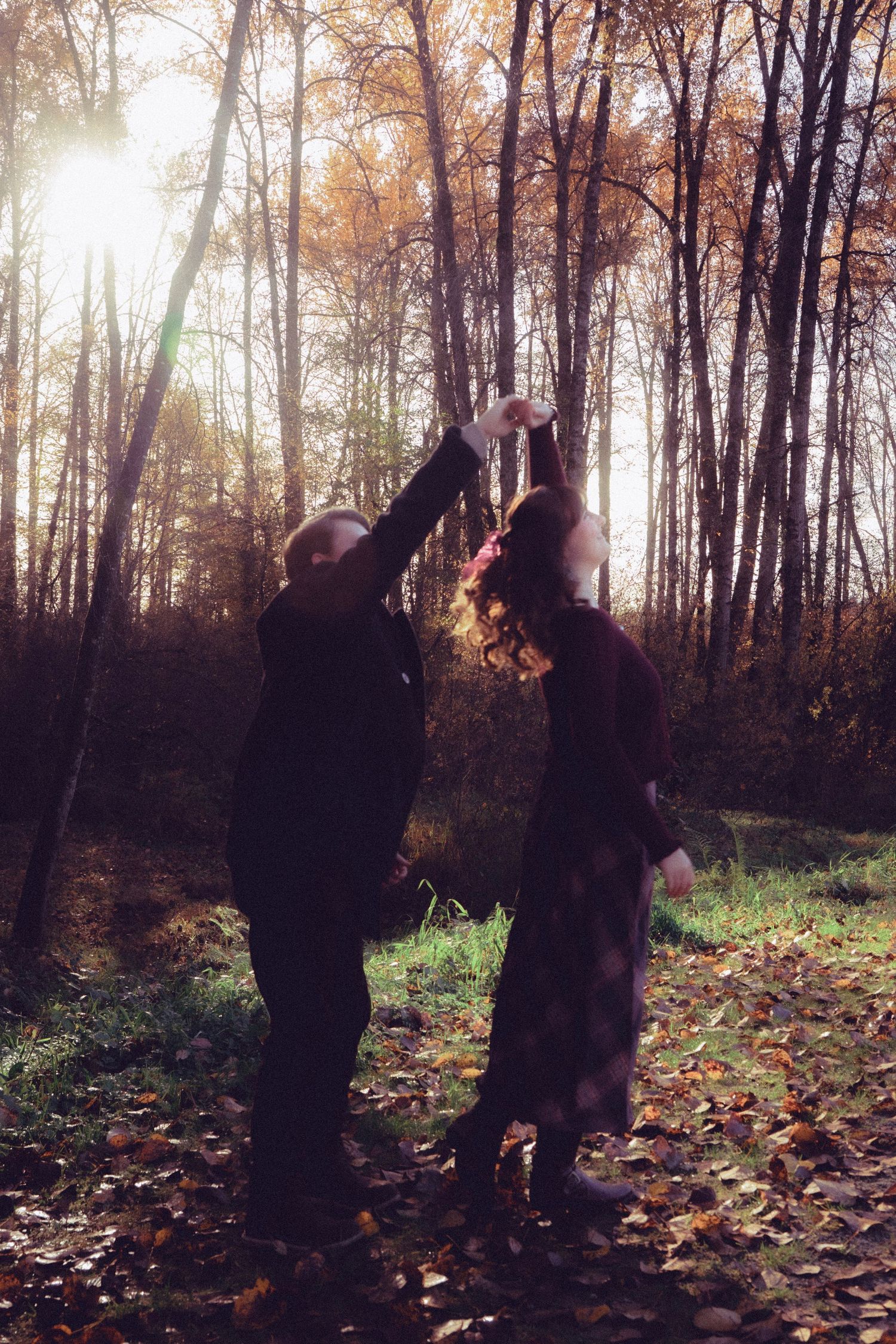 Couple dancing together in an autumn forest with golden sunlight filtering through bare trees and fallen leaves.