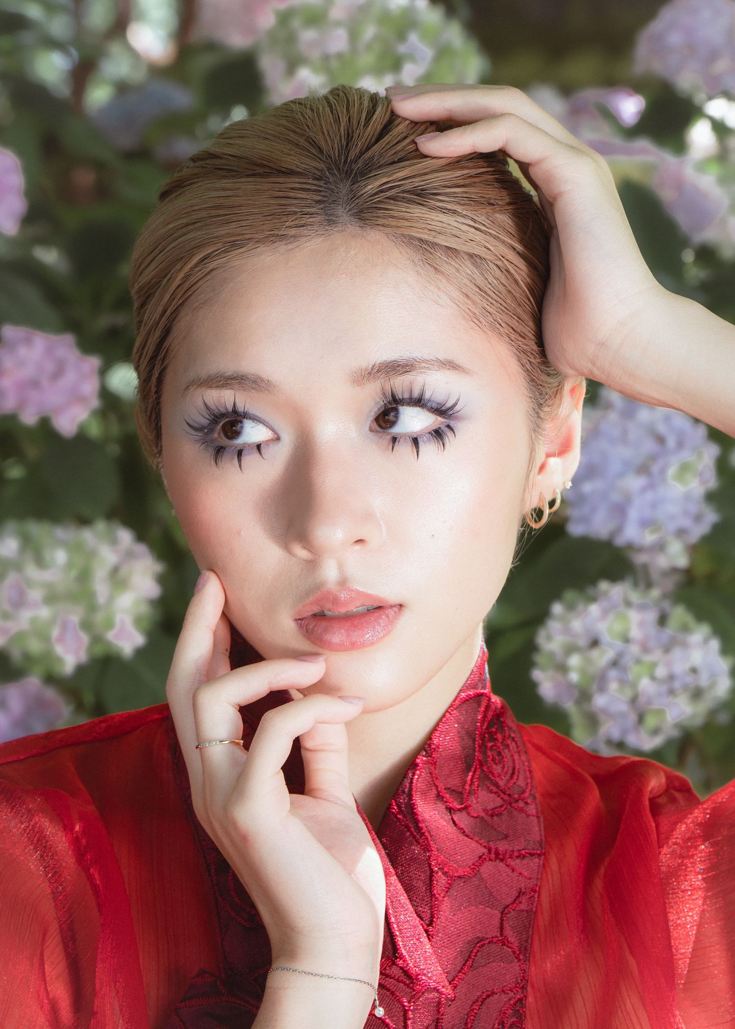 Young woman with dramatic purple eyelashes and bold eye makeup poses in red lace top among hydrangea flowers.