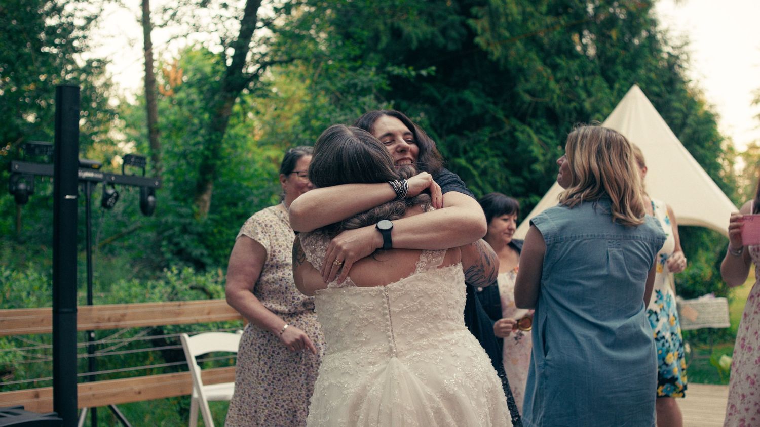 Bride in white wedding gown shares a warm embrace with guests outdoors at a garden wedding celebration.