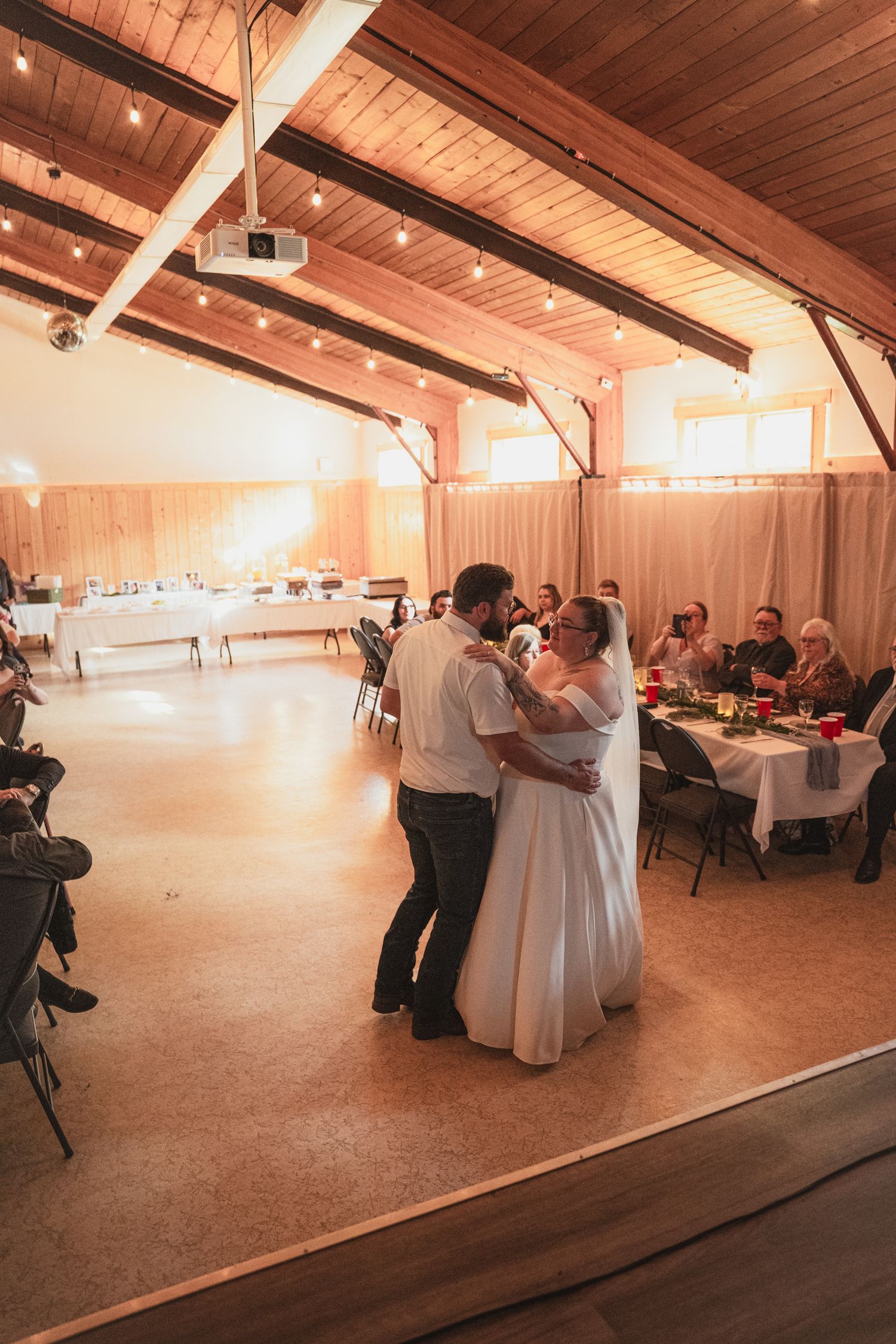 Newlyweds share a romantic first dance on the wooden floor of a warmly lit barn wedding reception.