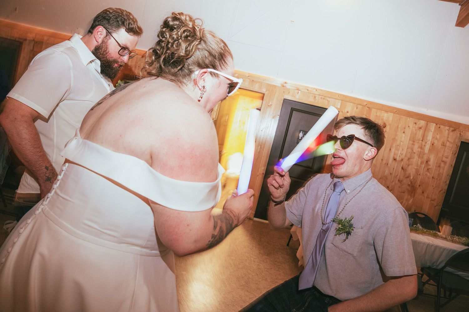 Wedding guests celebrate at reception, bride in white dress with groom and seated guest wearing novelty glasses holding glow stick.