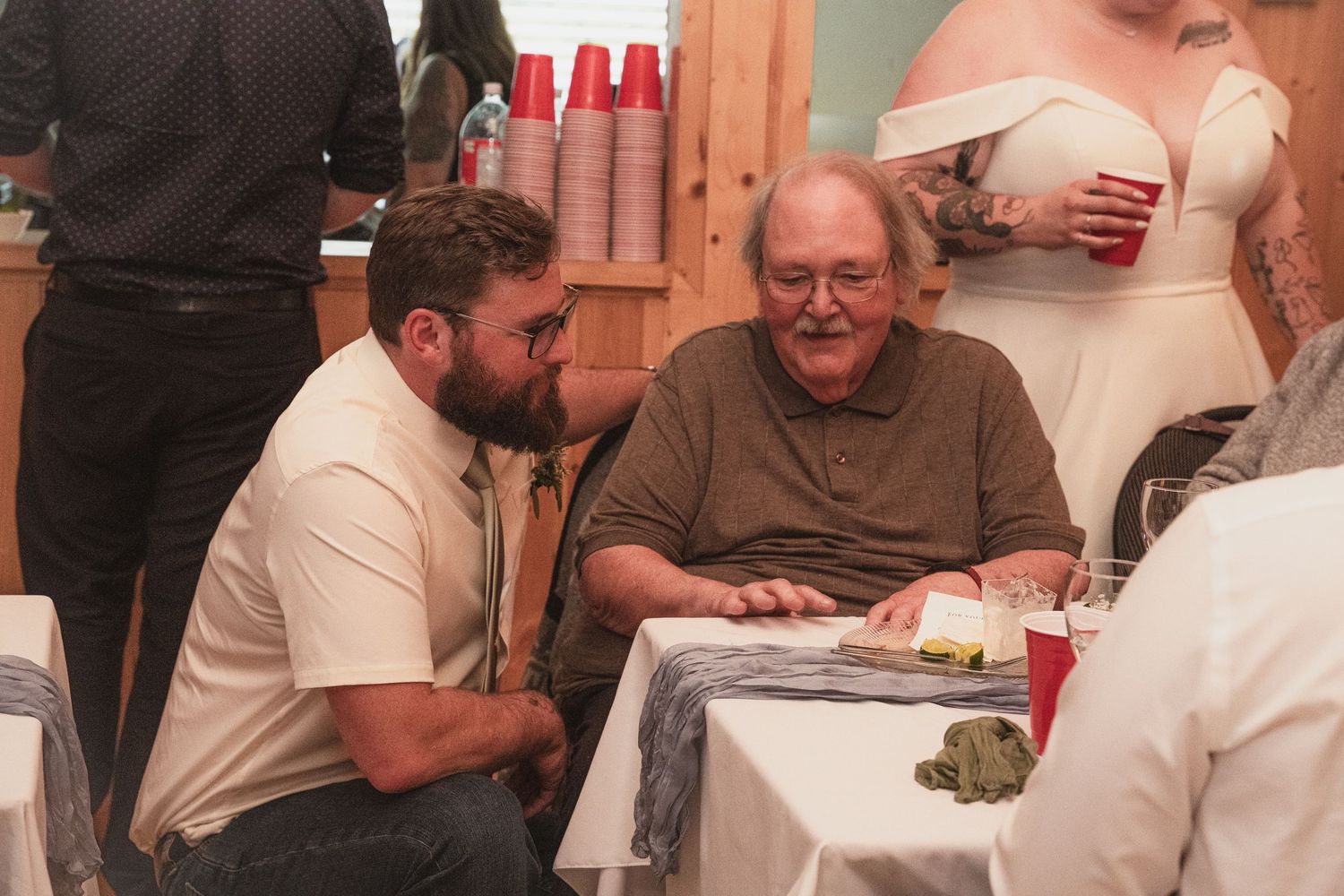 Young man kneels beside elderly guest at wedding reception table while bride stands nearby.