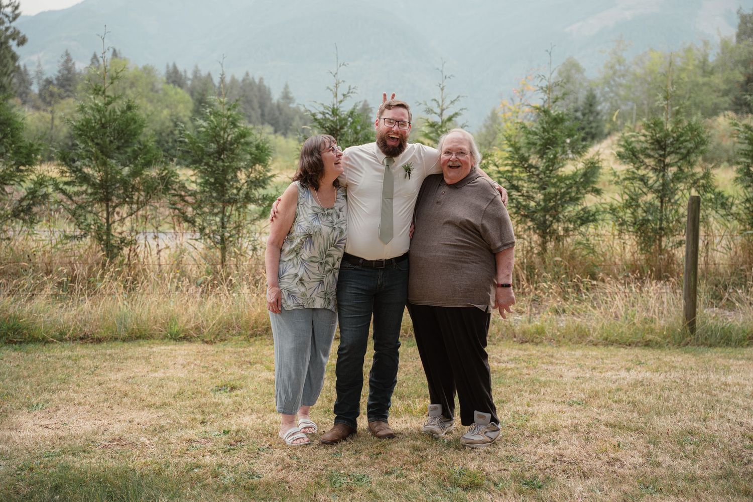 Groom in white shirt poses with two women smiling outdoors at a wedding venue.