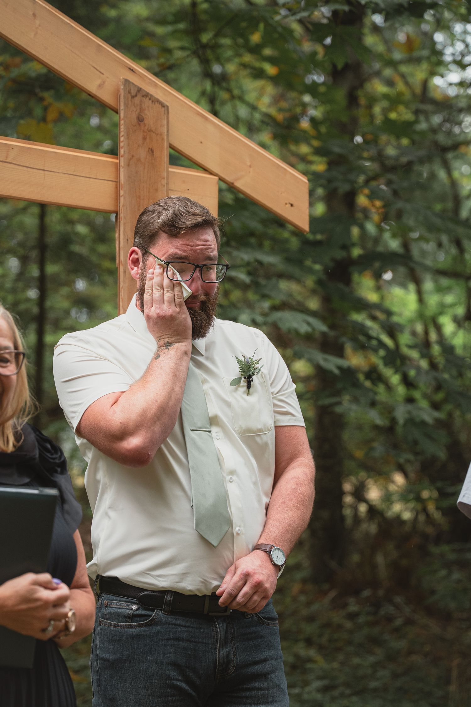 Emotional groom in white shirt and tie wipes tears during outdoor wedding ceremony beneath wooden arch.