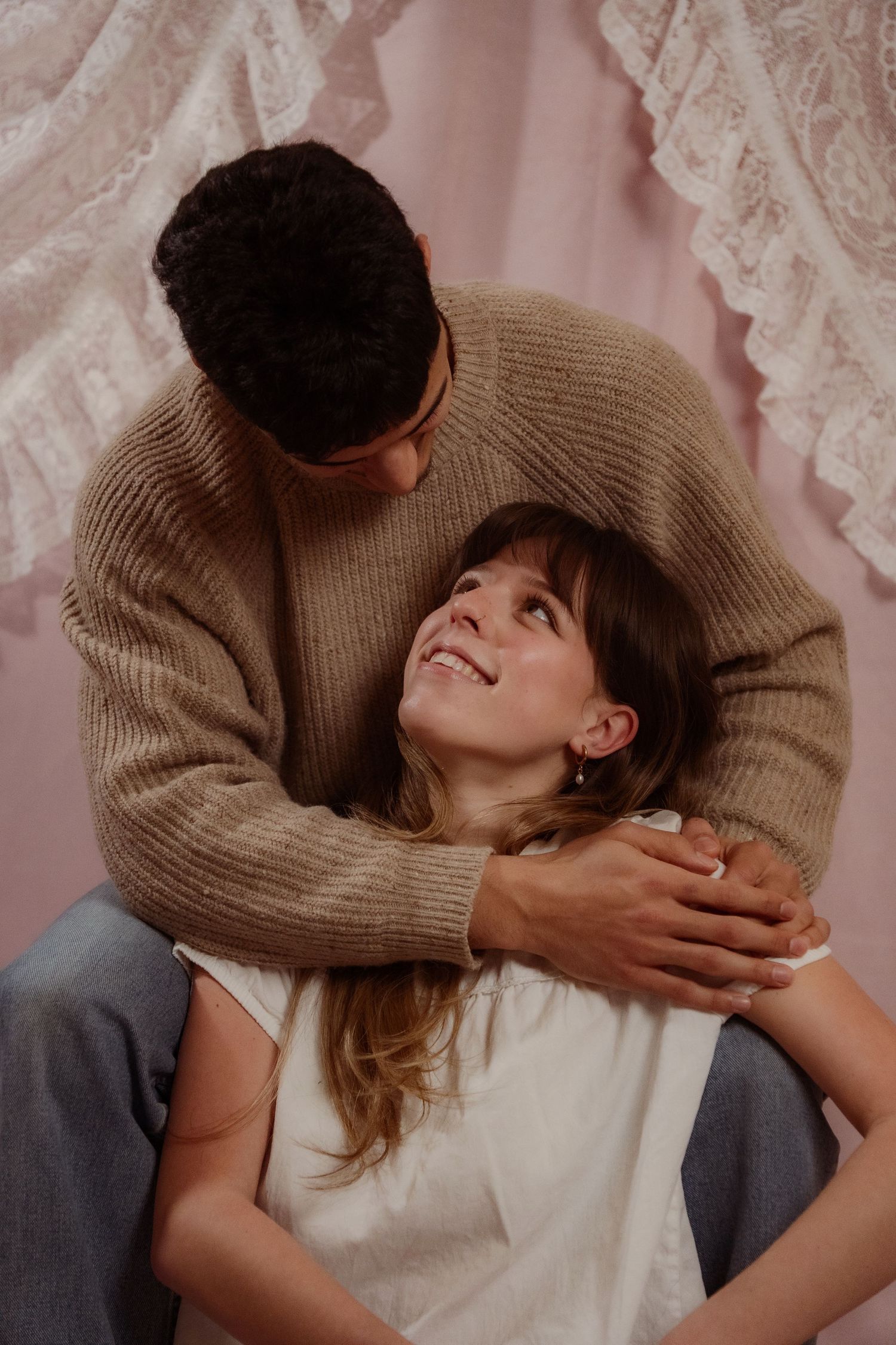 Young man in beige sweater tenderly embracing smiling woman from behind in a pink studio.