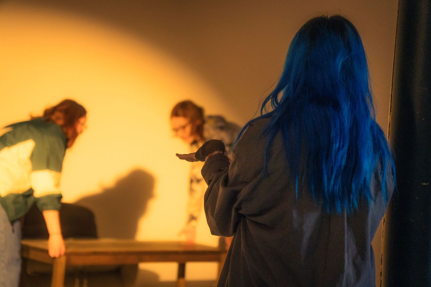 Blue-haired person holding a knife on a warmly lit film set with actors in the background.