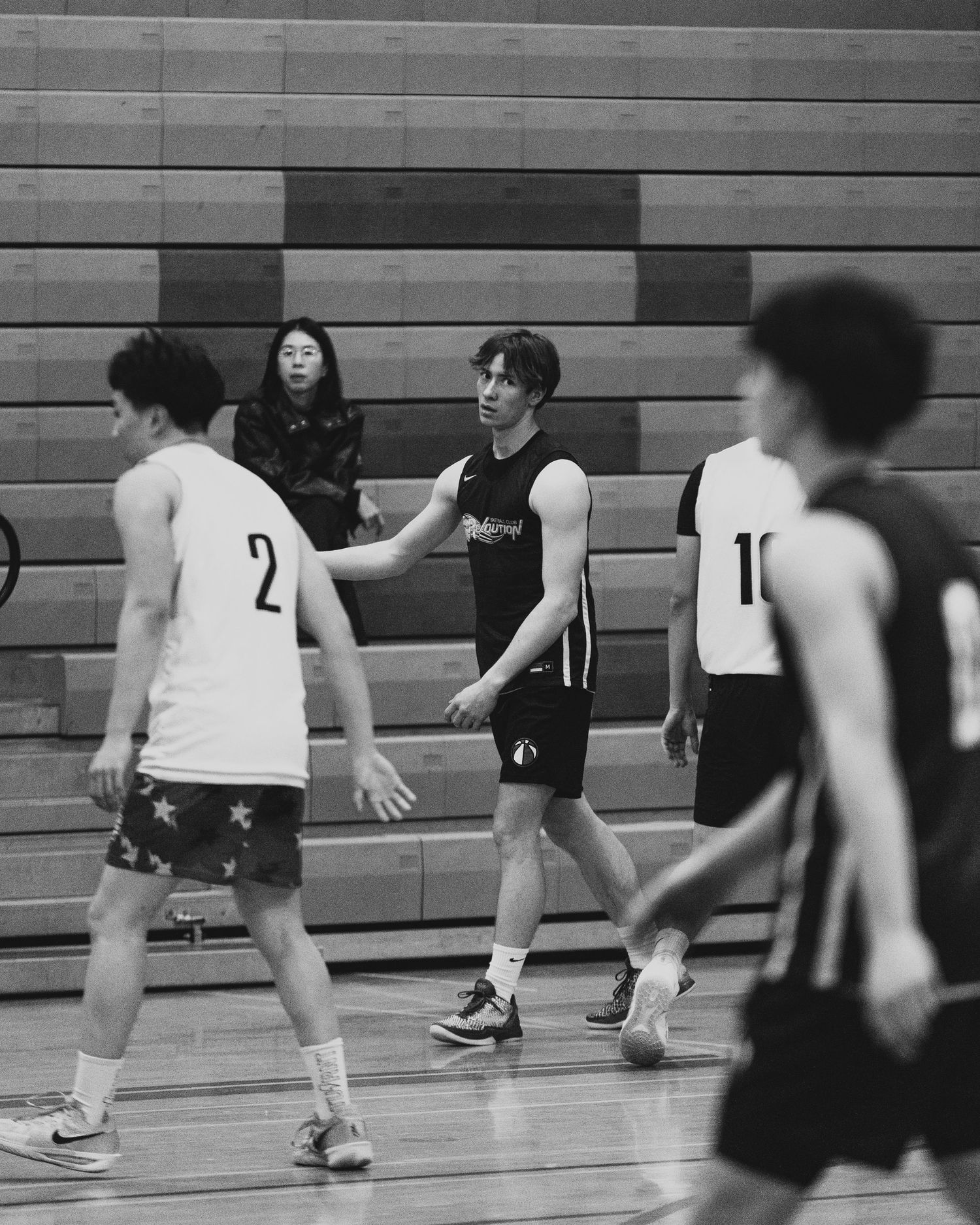 Black and white photo of basketball players on indoor court with spectators watching from bleachers in background.