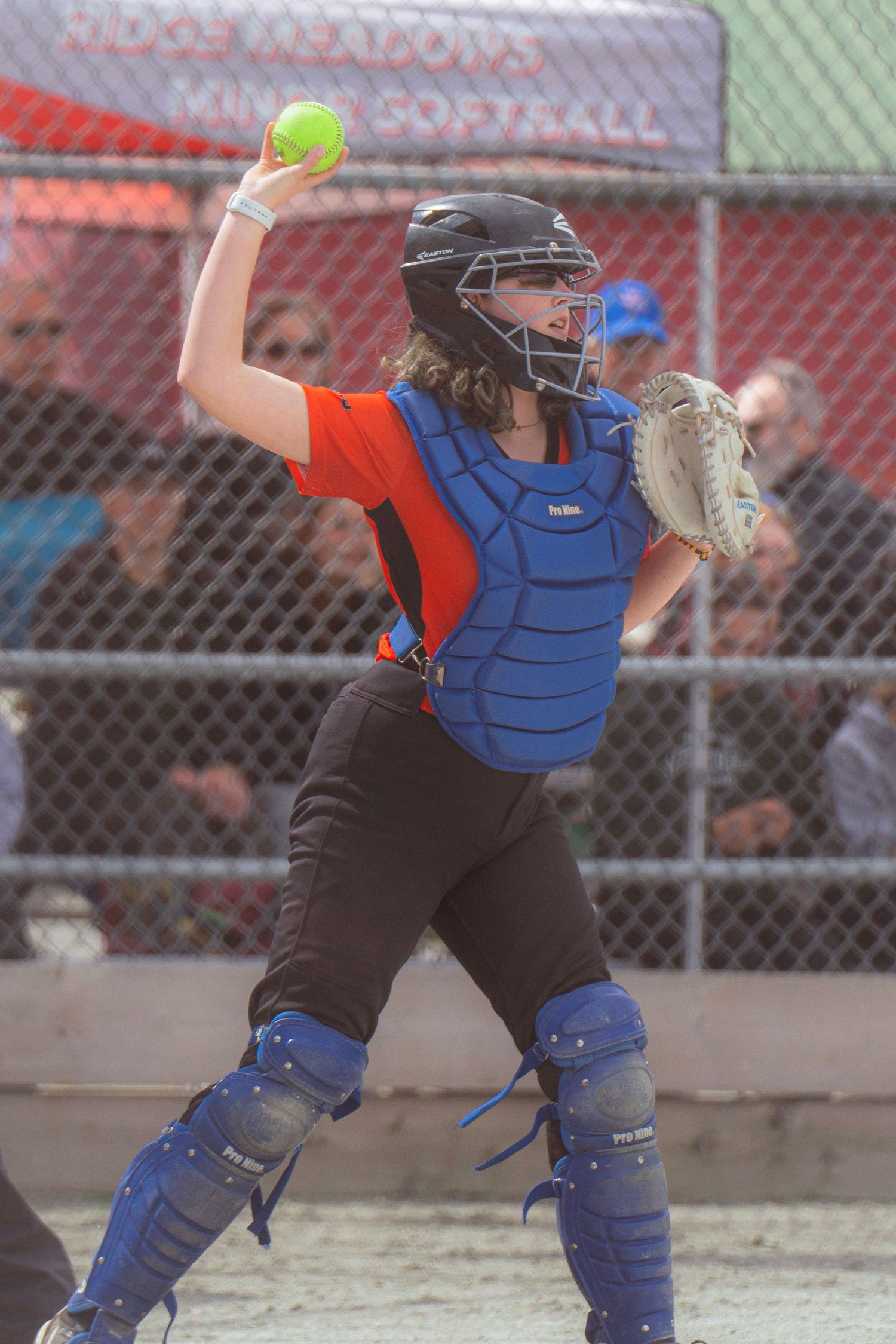 Softball catcher in orange jersey and blue gear winds up to throw yellow ball during game.