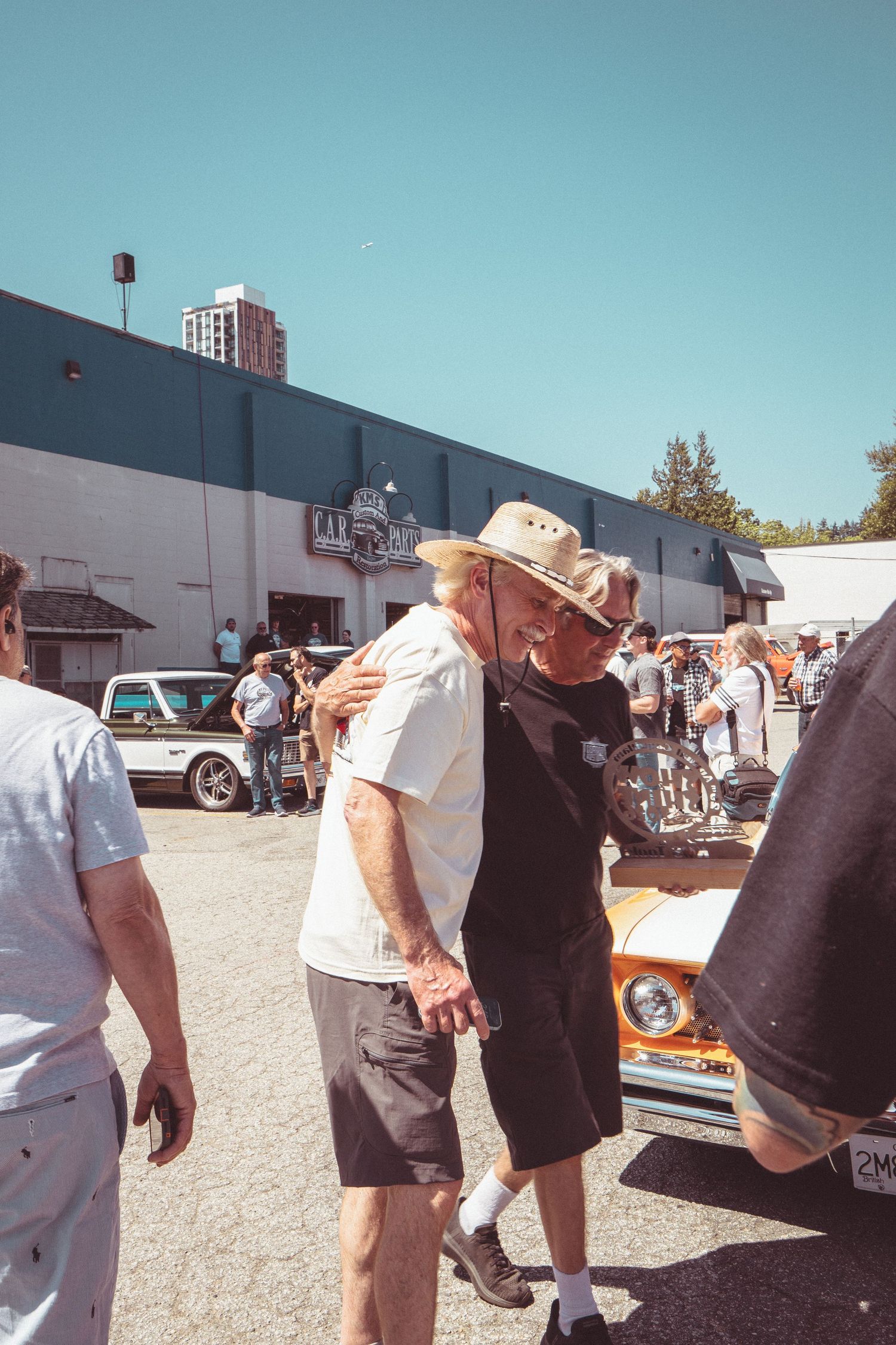 Two older men embracing at a sunny outdoor car show with classic vehicles in background.