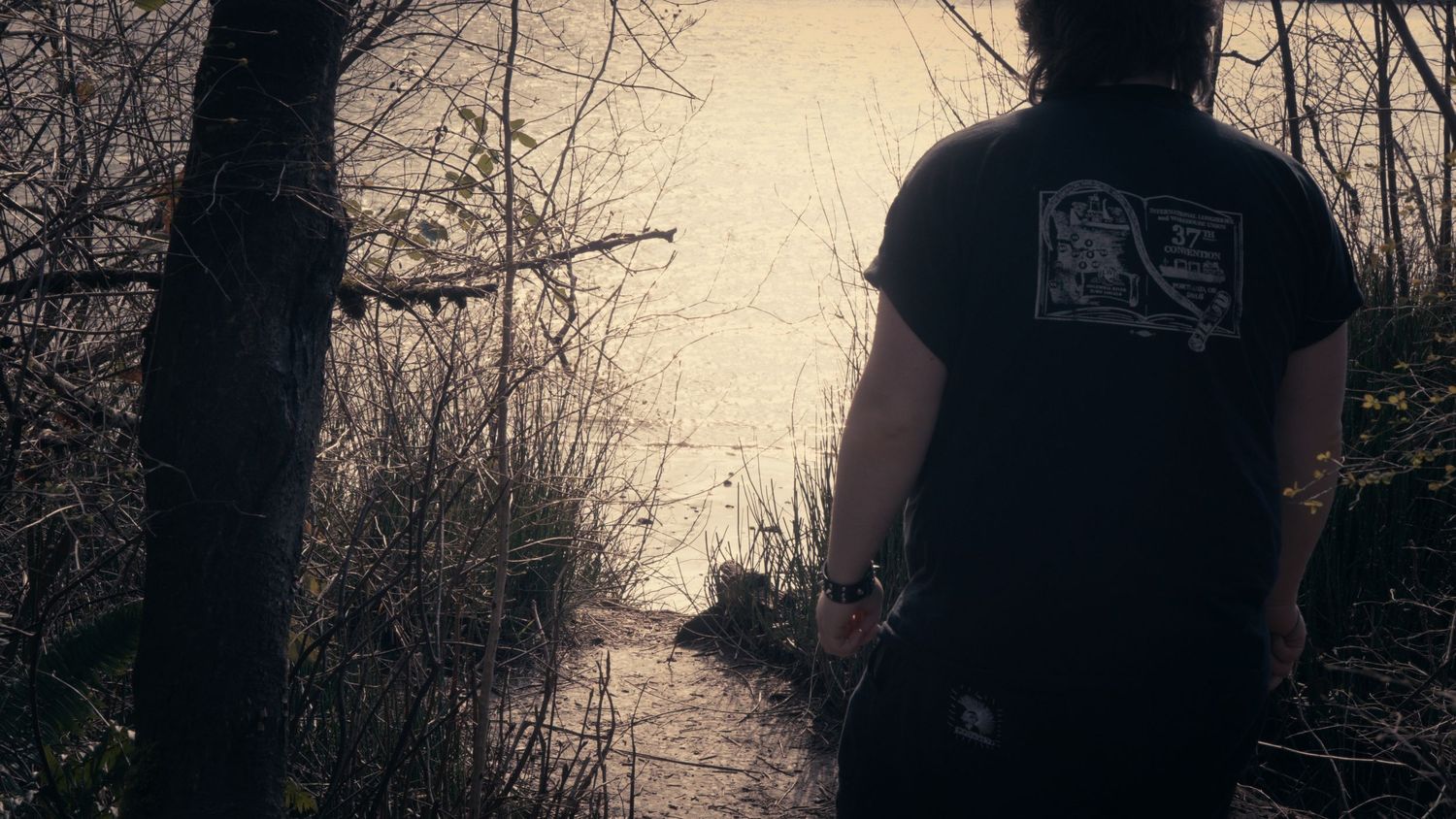 Person in black t-shirt standing by a reflective stream in woods.