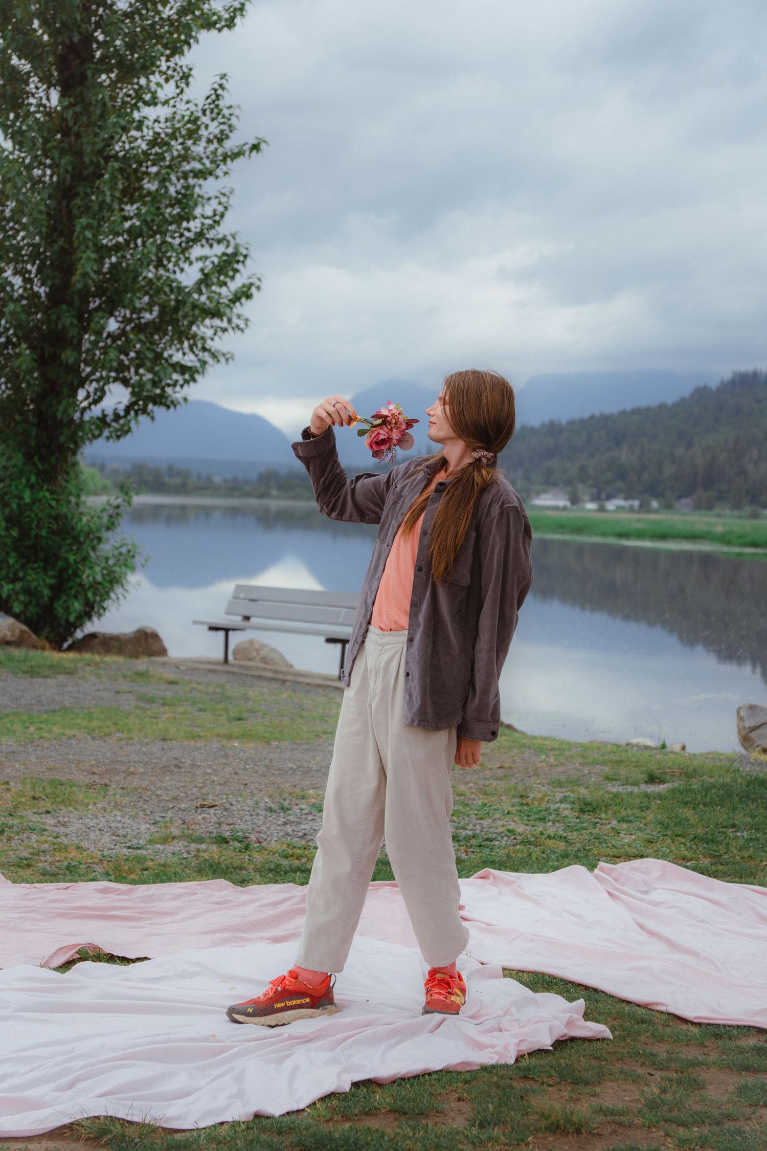 Young woman in casual outfit holding red flowers by a serene mountain lake on cloudy day.