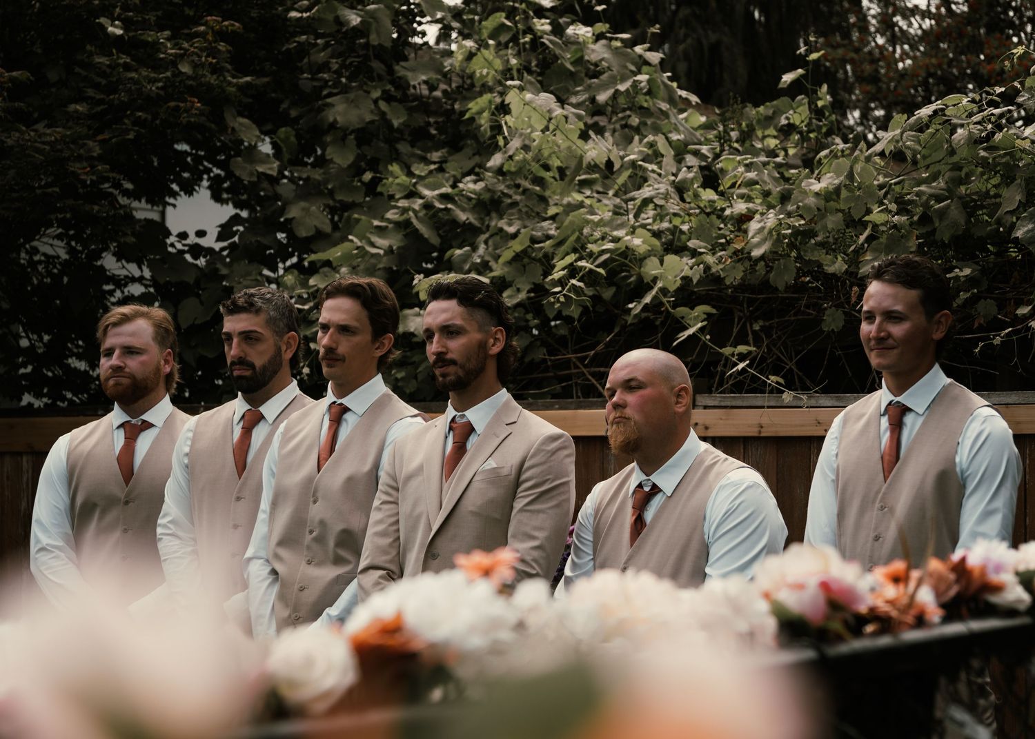 Six groomsmen in matching tan vests and rust ties stand in a row outdoors at a wedding ceremony.
