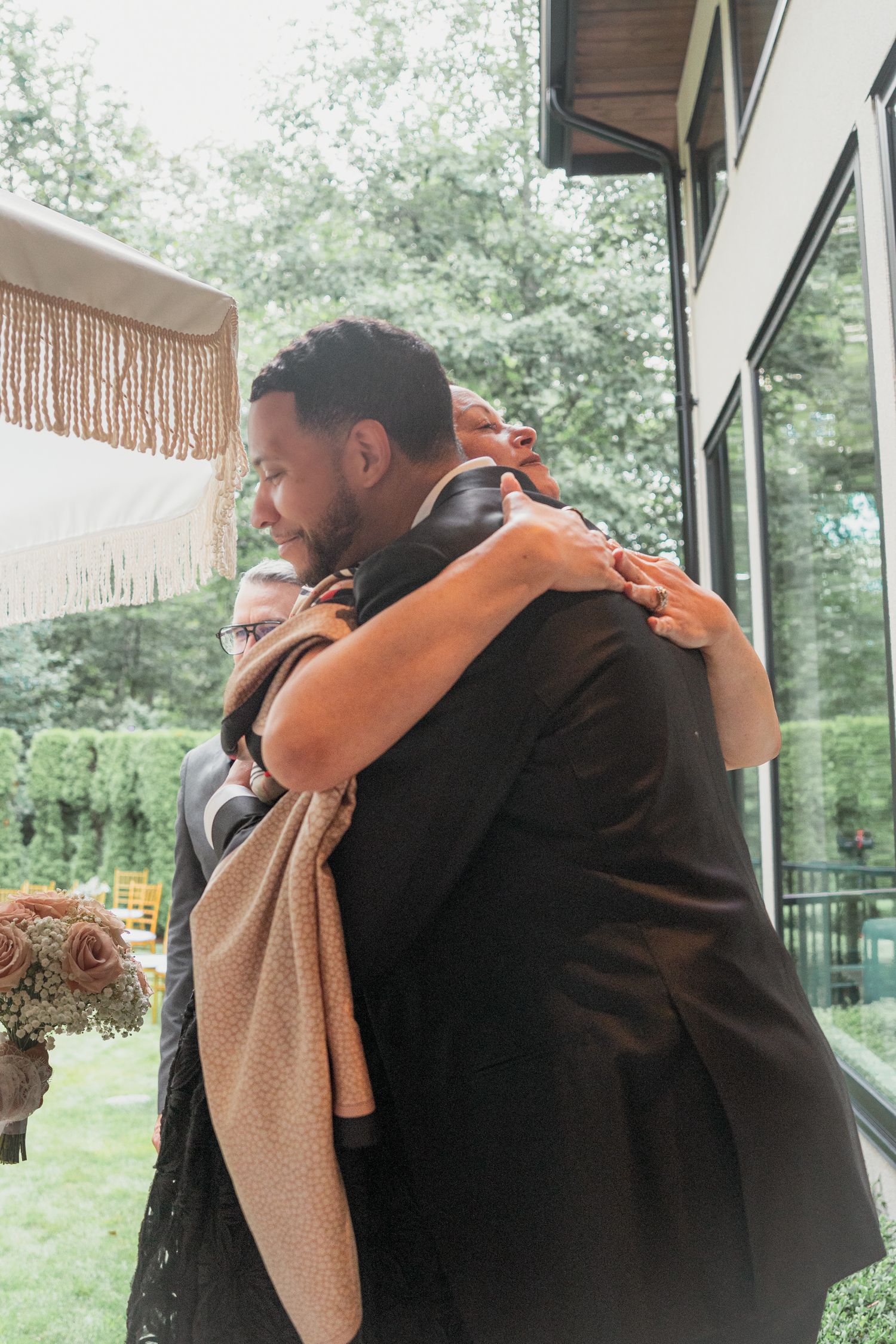 Two people share a warm embrace at an outdoor wedding ceremony, dressed in formal attire with greenery in the background.