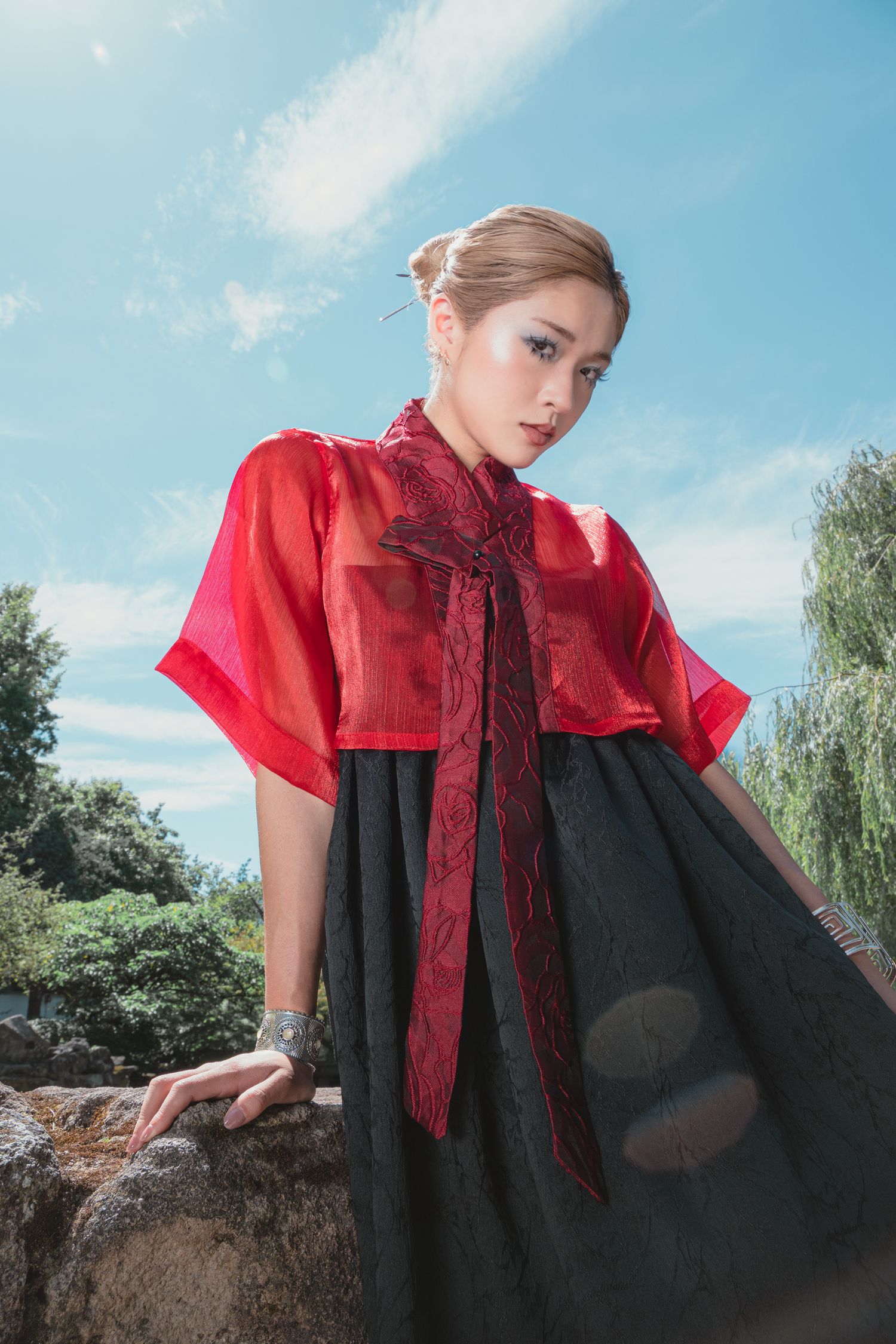 Young woman in red organza top and black skirt posing outdoors against blue sky in traditional Asian-inspired fashion.